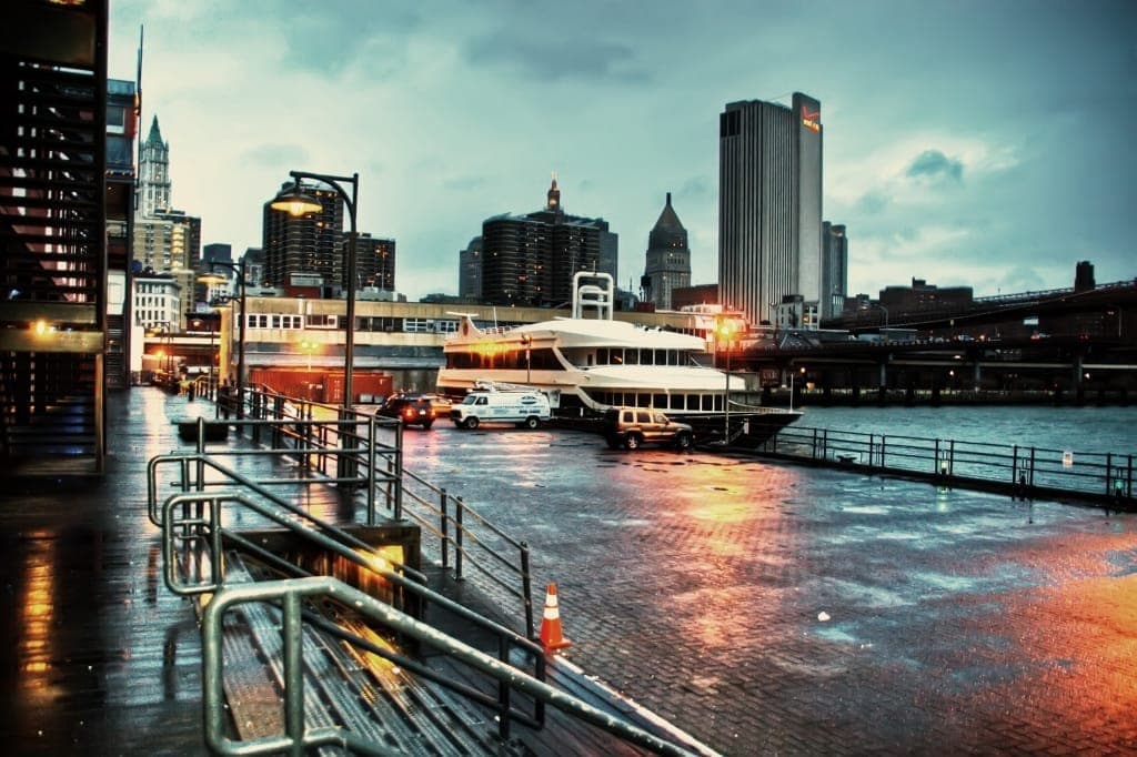 ท่าเรือหินกรวดเปียกในย่าน South Street Seaport Historic District พร้อมเรือที่จอดเทียบท่า รั้วริมน้ำ และตึกระฟ้าของแมนฮัตตันในแสงยามเย็น