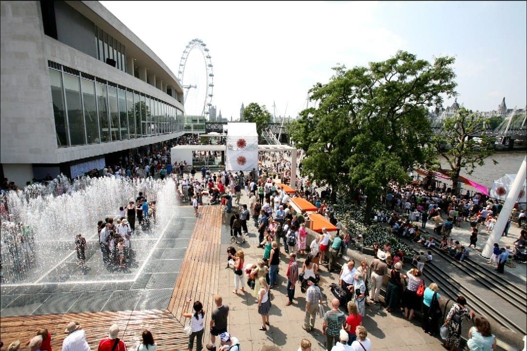An einem sonnigen Tag versammeln sich Menschenmengen um die Brunnen vor dem Southbank Centre, im Hintergrund sind das London Eye und die Themse zu sehen.