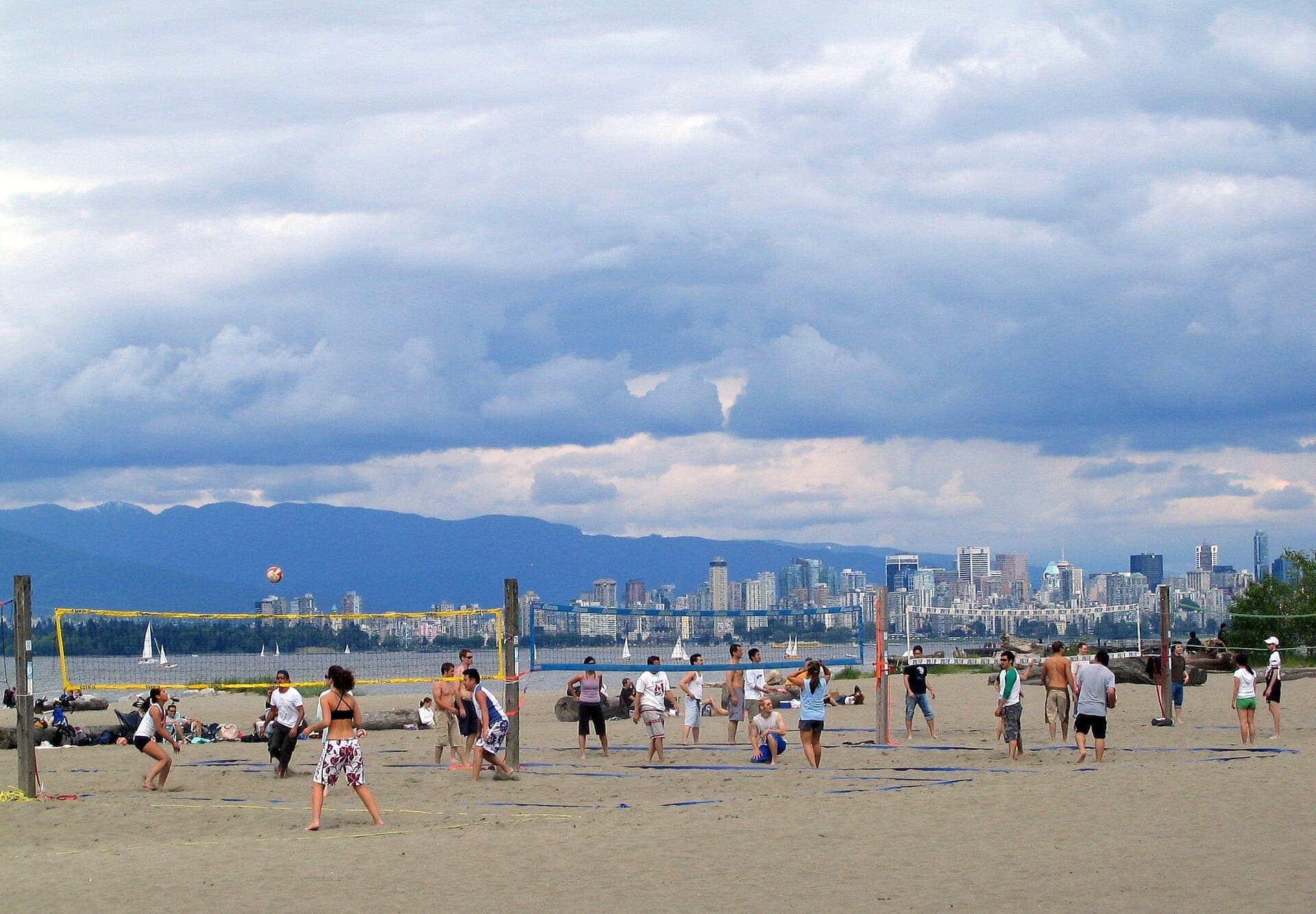 กลุ่มคนเล่นวอลเลย์บอลชายหาดบน Spanish Banks Beach ที่เต็มไปด้วยทราย โดยมีตัวเมือง Vancouver เรือใบ และภูเขาเป็นฉากหลัง