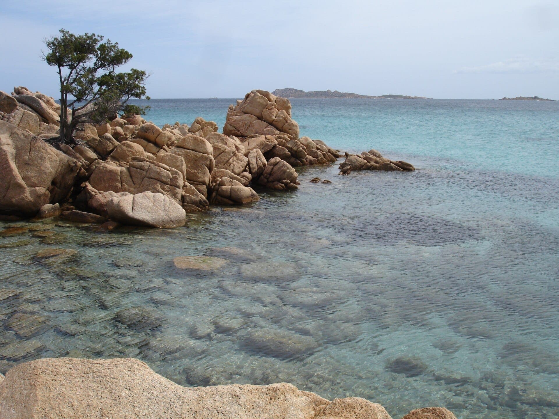 Klares türkisfarbenes Wasser und abgerundete Granitfelsen an der Spiaggia di Capriccioli, mit einem einzelnen Baum und fernen Inseln unter einem strahlenden Himmel.