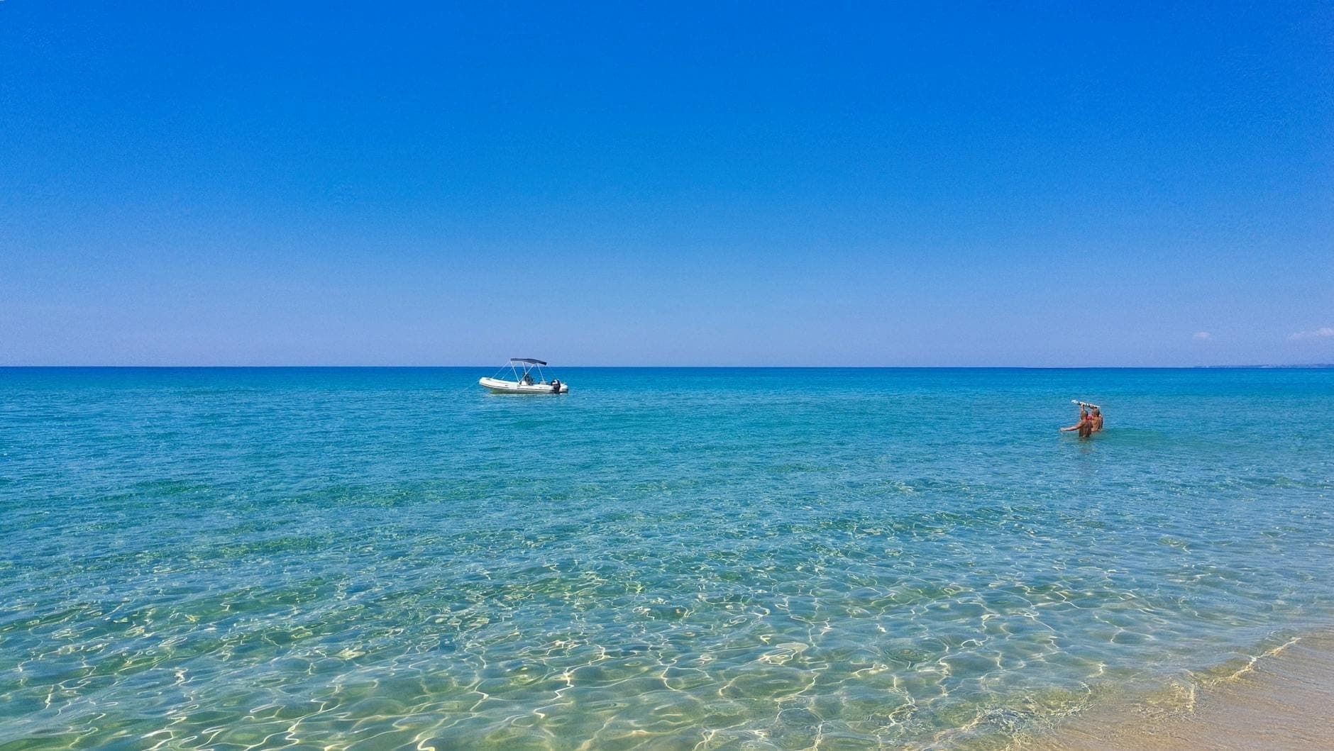Weiter Blick auf einen ruhigen Strand mit klarem türkisfarbenem Wasser, einem kleinen Boot und einem Schwimmer unter strahlend blauem Himmel.