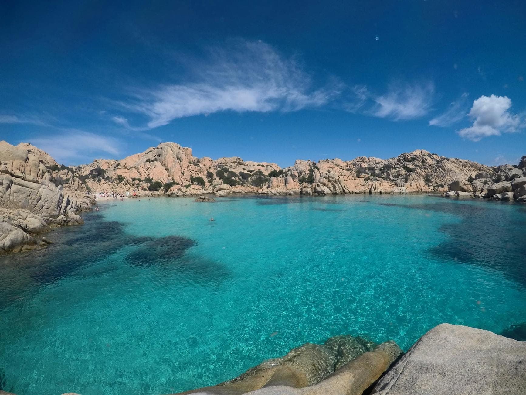 Türkisfarbenes Wasser umgeben von rosafarbenem Granit unter blauem Himmel – die abgeschiedene Doppelbucht der Cala Coticcio auf Caprera.