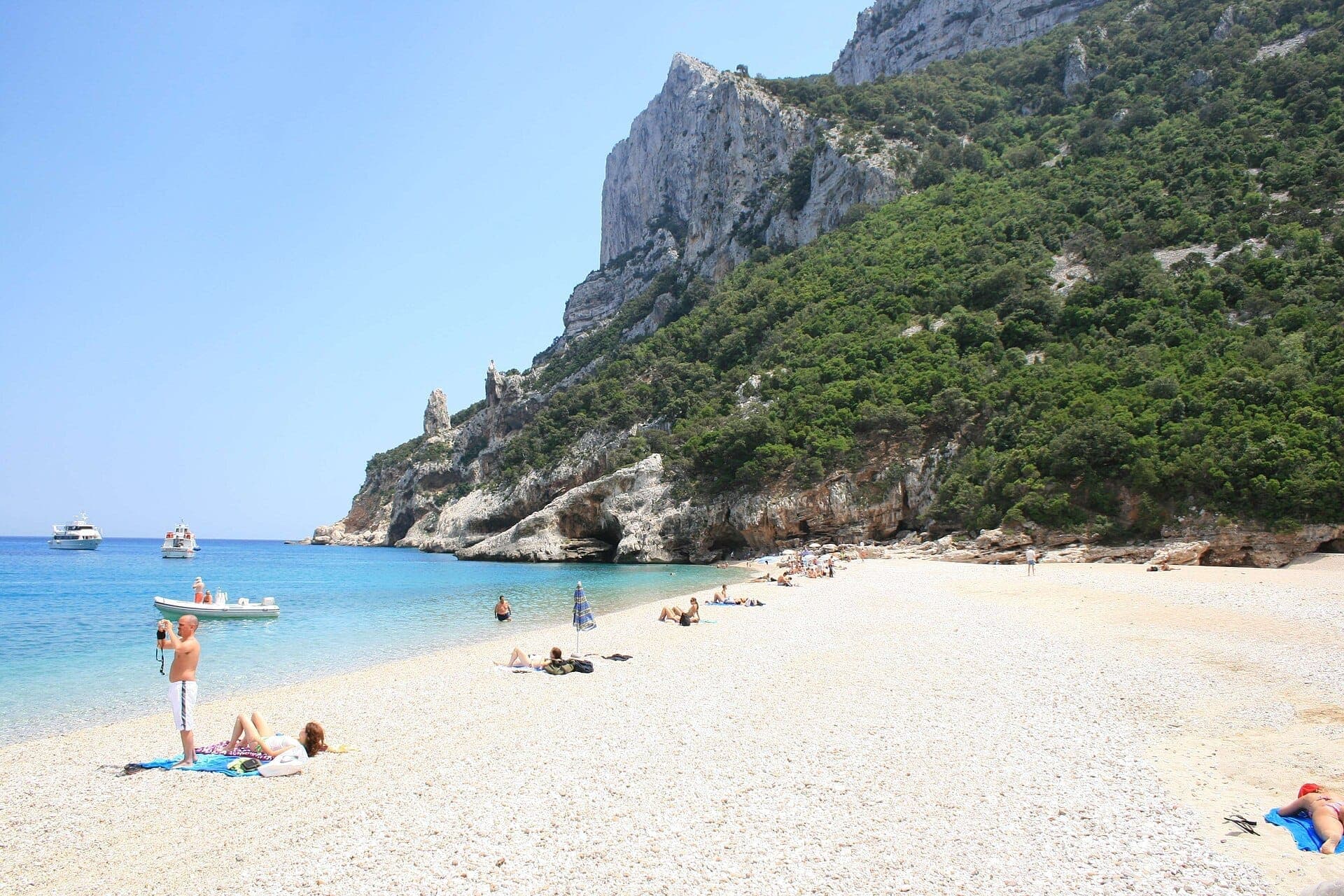 Breiter Kiesstrand an der Cala Sisine mit türkisfarbenem Wasser, Sonnenbadenden, vor Anker liegenden Booten und beeindruckenden Kalksteinklippen mit üppiger Vegetation.
