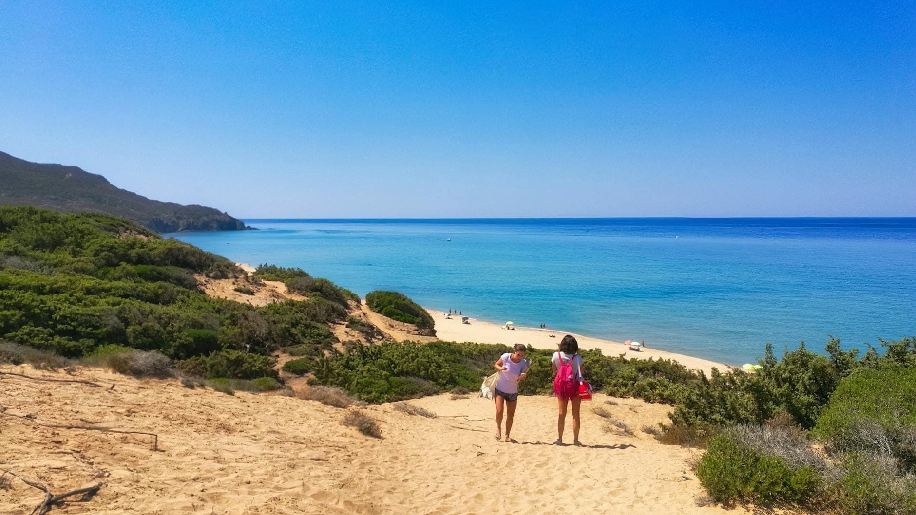 Zwei Menschen gehen sandige Dünen hinunter zu einem unberührten Strand mit klarem türkisfarbenem Wasser, sattgrünen Büschen und blauem Himmel an einem sonnigen Tag.