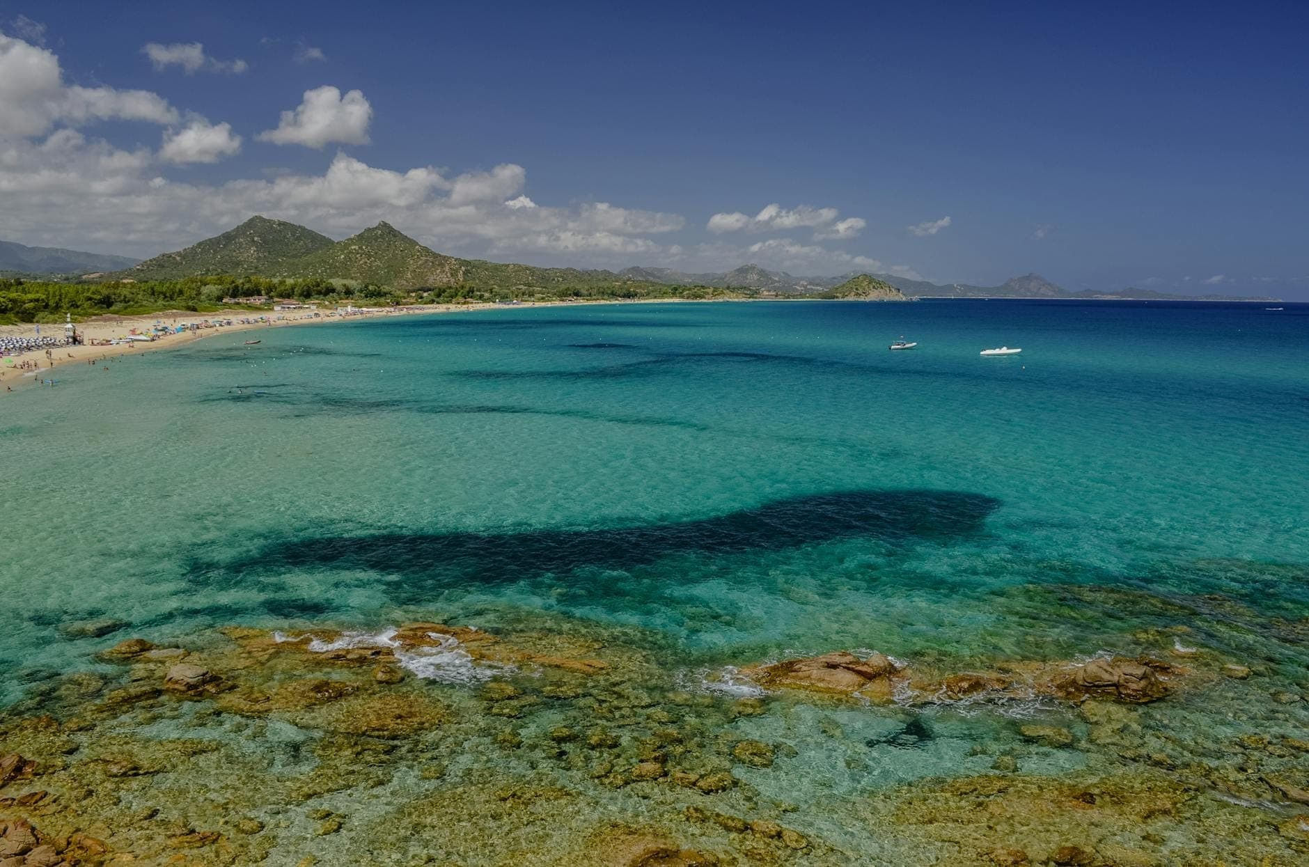 Weitblick über die Spiaggia di Costa Rei mit klarem türkisblauem Wasser, felsigem Vordergrund, weißem Sandstrand und sanften Hügeln unter blauem Himmel.