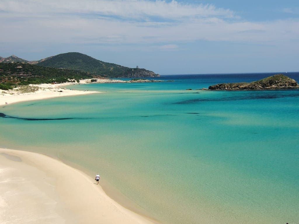 Der weite Bogen aus hellem Sand und türkisfarbenem Wasser der Spiaggia di Tuerredda, mit grünen Hügeln und einem kleinen Felseninselchen im Hintergrund.