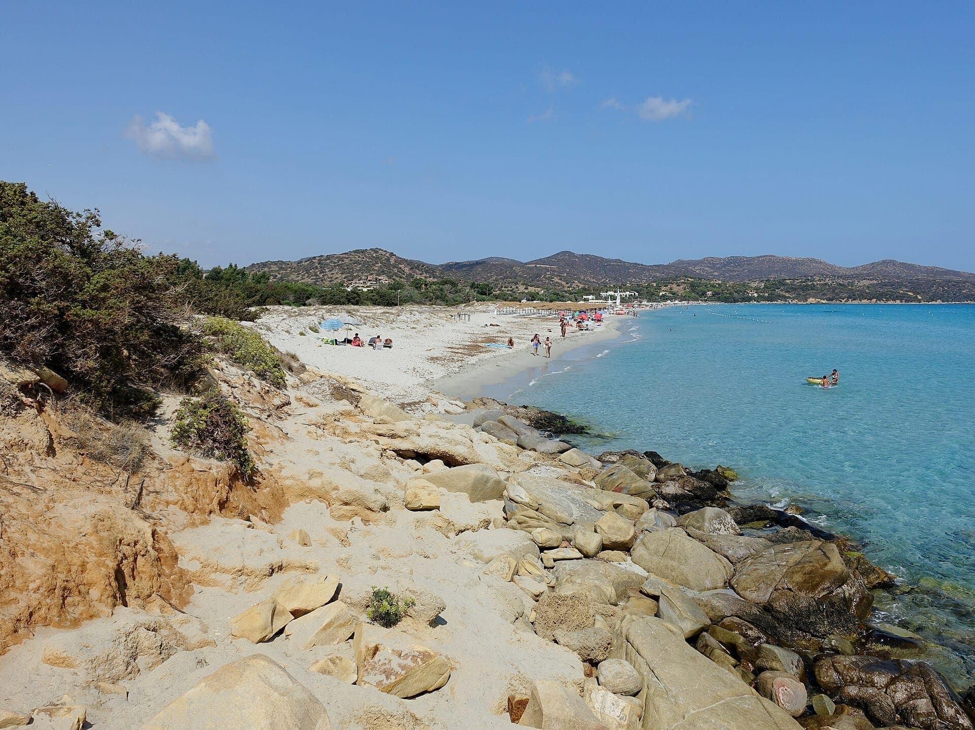 La Spiaggia di Simius tiene arena blanca y suave, agua turquesa, orilla rocosa, bañistas y colinas bajo un cielo azul despejado en Cerdeña.