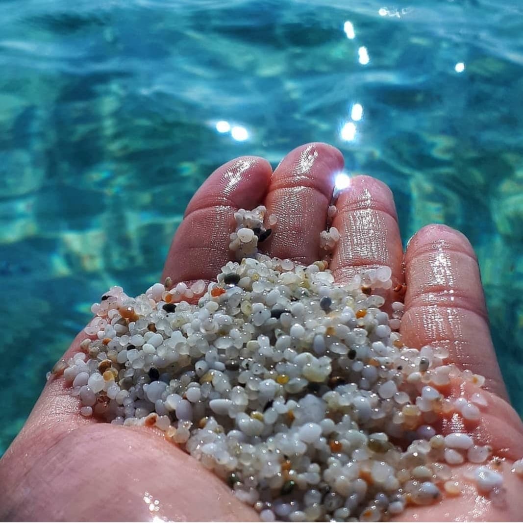 Nahaufnahme einer Hand, die bunte Quarzkörner an der Spiaggia di Is Arutas hält, mit klarem blauem Wasser, das im Hintergrund glitzert.