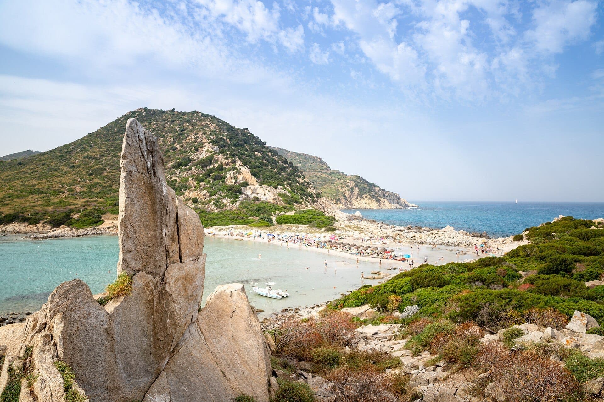 Weitblick auf Spiaggia di Punta Molentis mit zerklüftetem Granitfelsen, türkisfarbenem Wasser, grünen Hügeln und bunten Sonnenschirmen unter einem teilweise bewölkten Himmel.