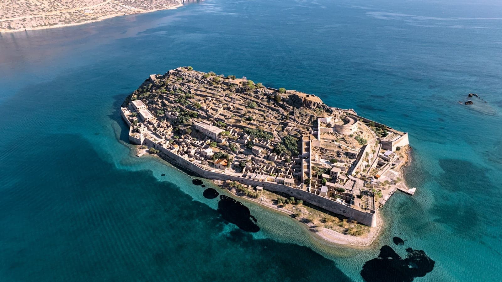 Luftaufnahme der Insel Spinalonga, umgeben von türkisblauem Wasser, mit der gesamten Festungsanlage, detaillierten Steinmauern und Ruinen vor dem Hintergrund des blauen Meeres und der kretischen Küste.