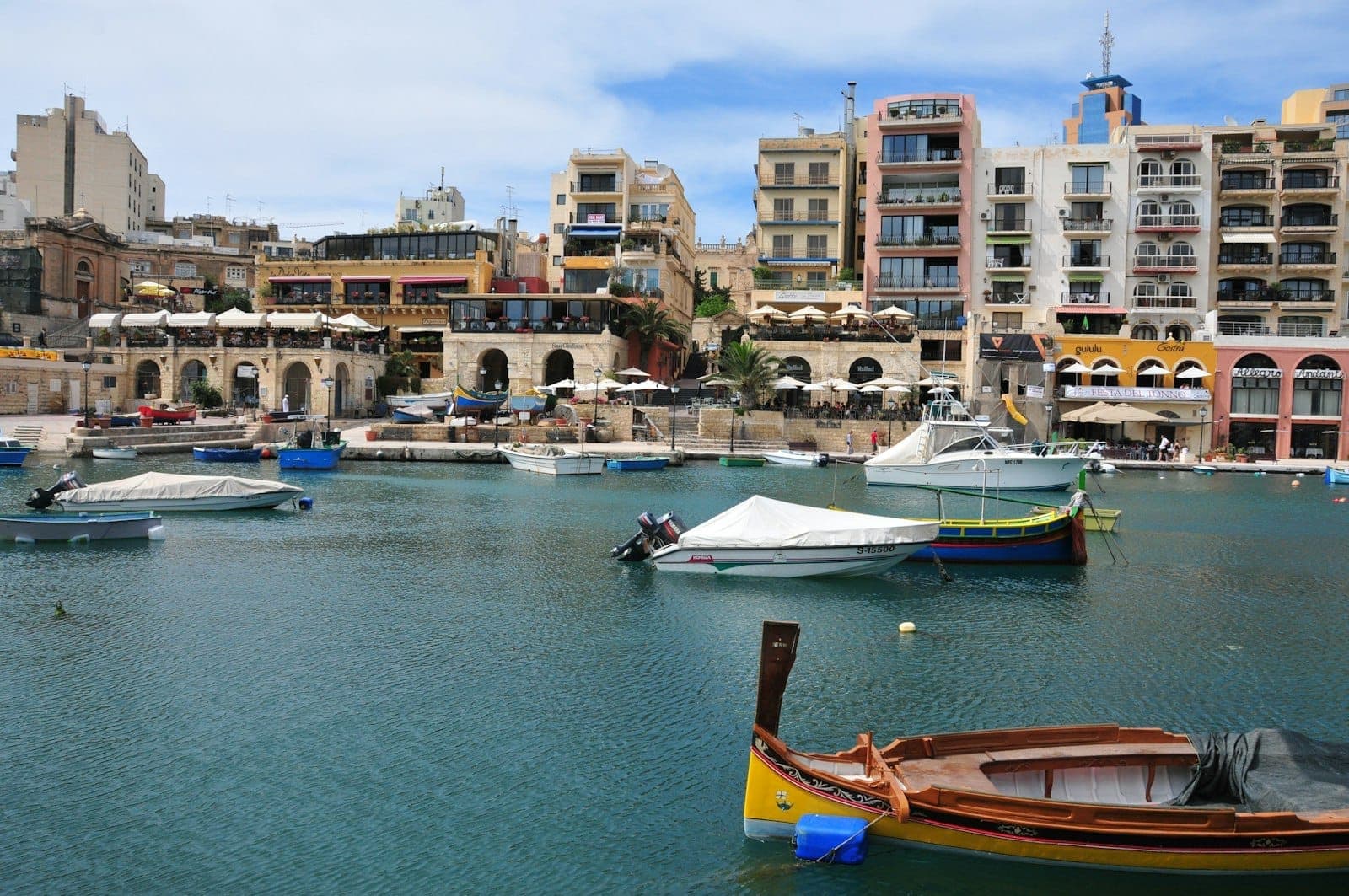 Traditionelle Fischerboote schwimmen in der Spinola Bay, Malta, mit bunten Gebäuden, Restaurantterrassen und blauem Wasser unter einem teilweise bewölkten Himmel.