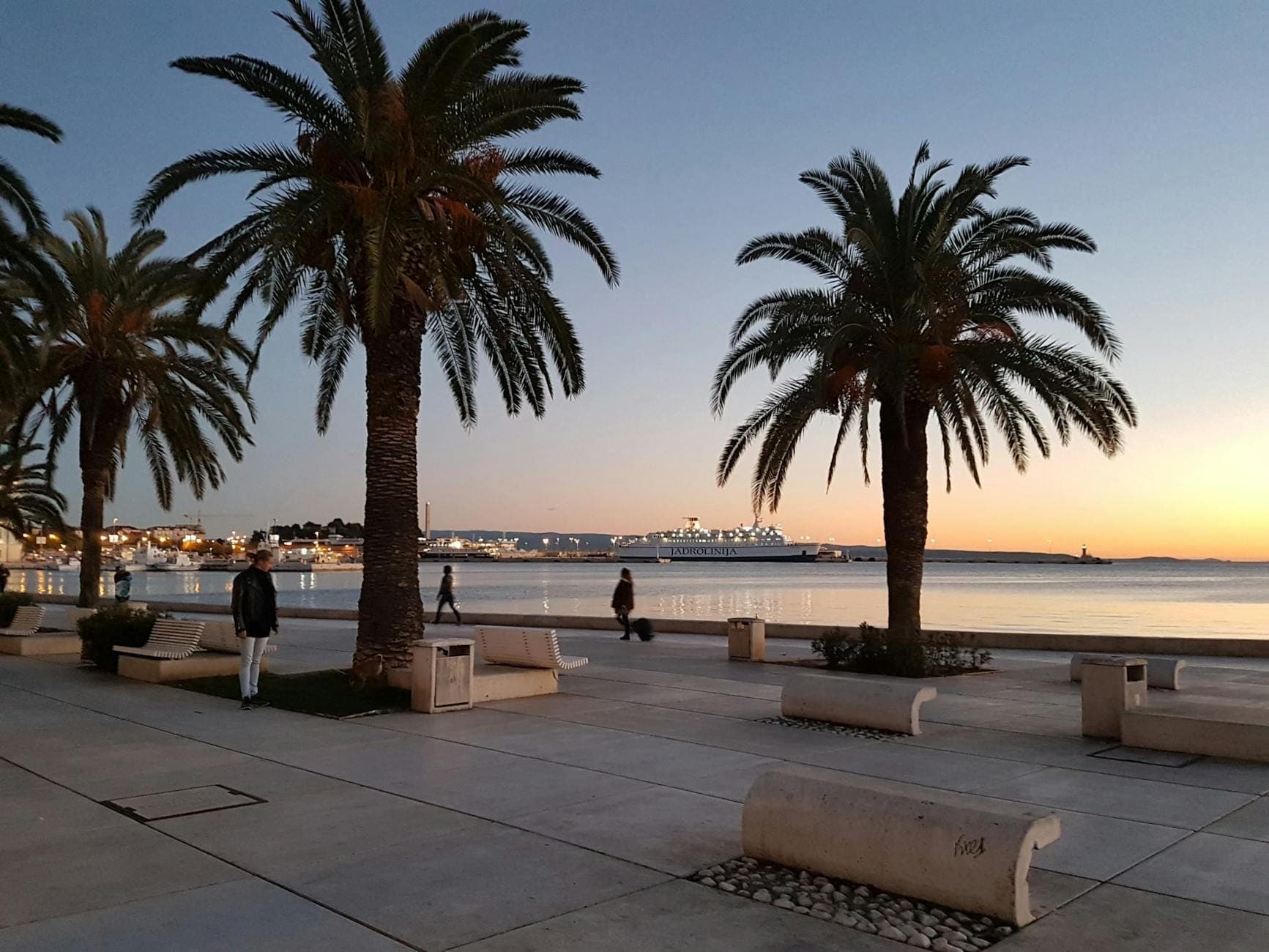 Palme e panchine fiancheggiano il lungomare Riva a Spalato al tramonto, con persone che passeggiano e il porto sullo sfondo.