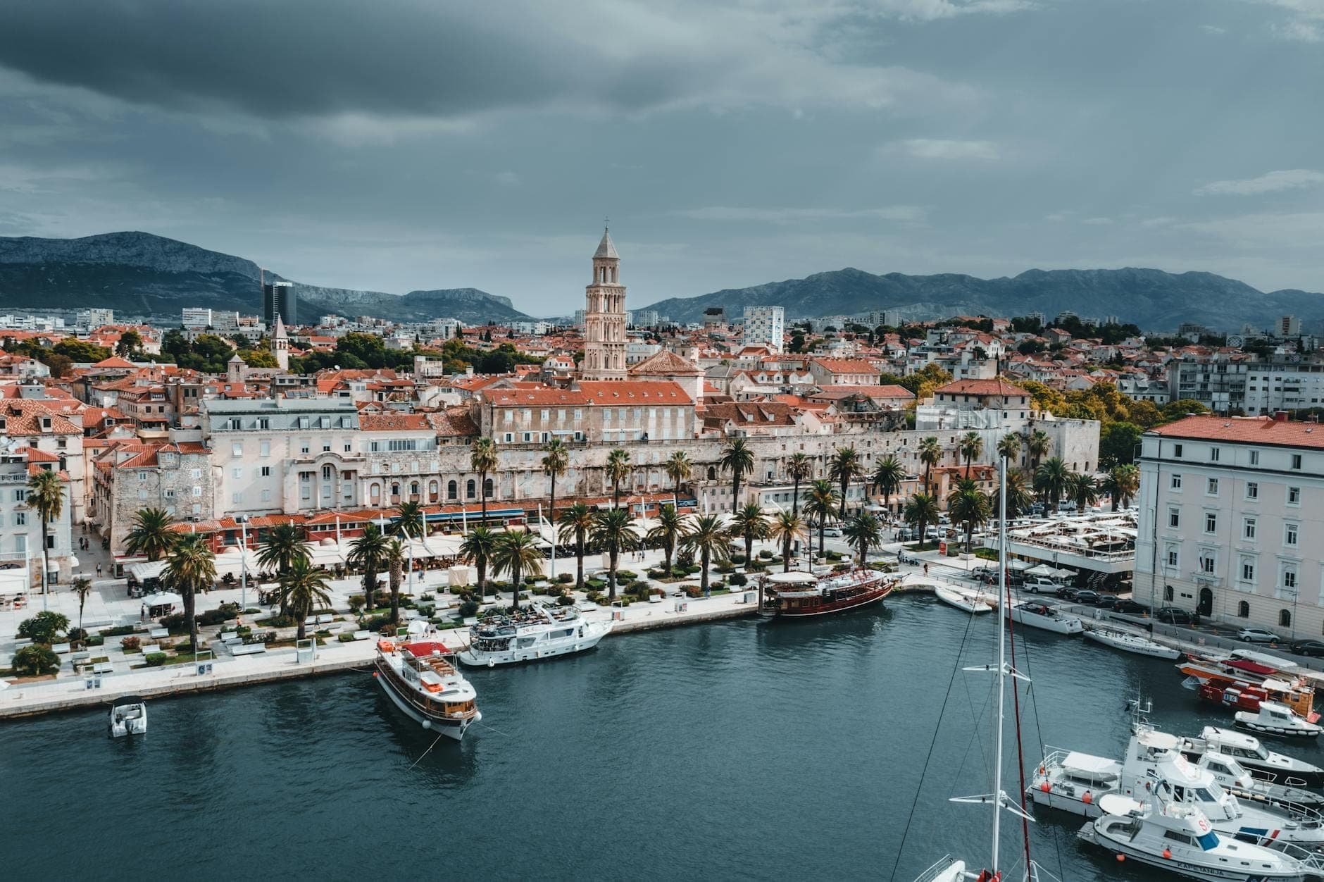 Vista panoramica del lungomare di Split con il Palazzo di Diocleziano, il viale alberato di palme, barche ormeggiate e montagne spettacolari sotto un cielo nuvoloso.