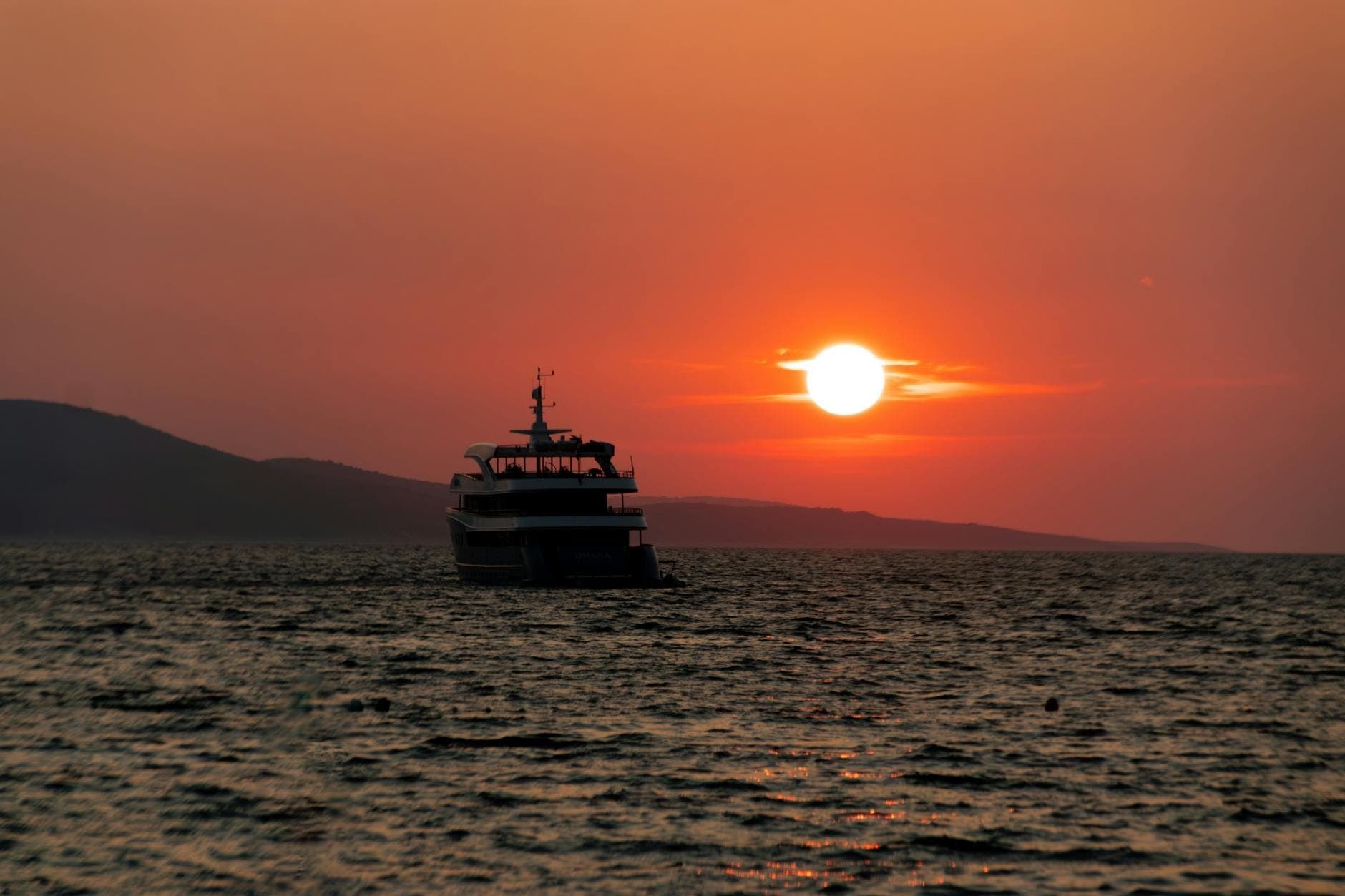 Silhouette d'un bateau sur un coucher de soleil spectaculaire au-dessus de la mer, avec un littoral vallonné en arrière-plan et un ciel orange.