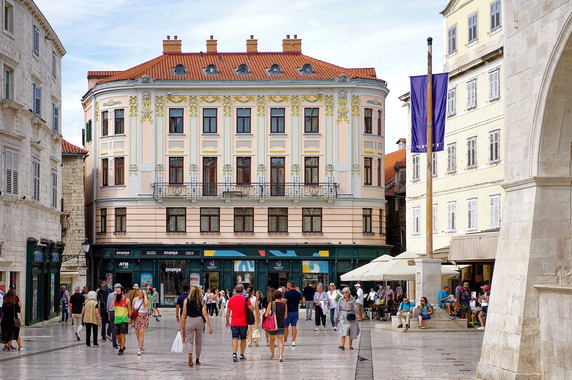 Gruppi di persone passeggiano e si rilassano nella storica piazza principale di Split, circondata da ornati edifici color pastello con tetti rossi sotto un cielo luminoso.