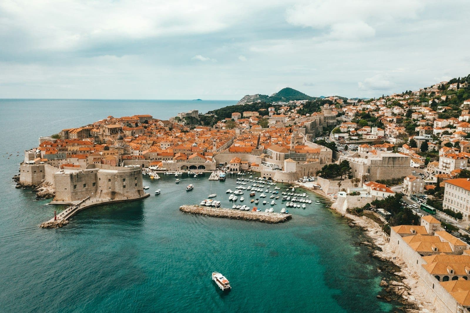 Vista aerea della città vecchia di Dubrovnik con i tetti arancioni, le mura, il porto e una piccola barca nelle acque turchesi dell'Adriatico sotto un cielo parzialmente nuvoloso.