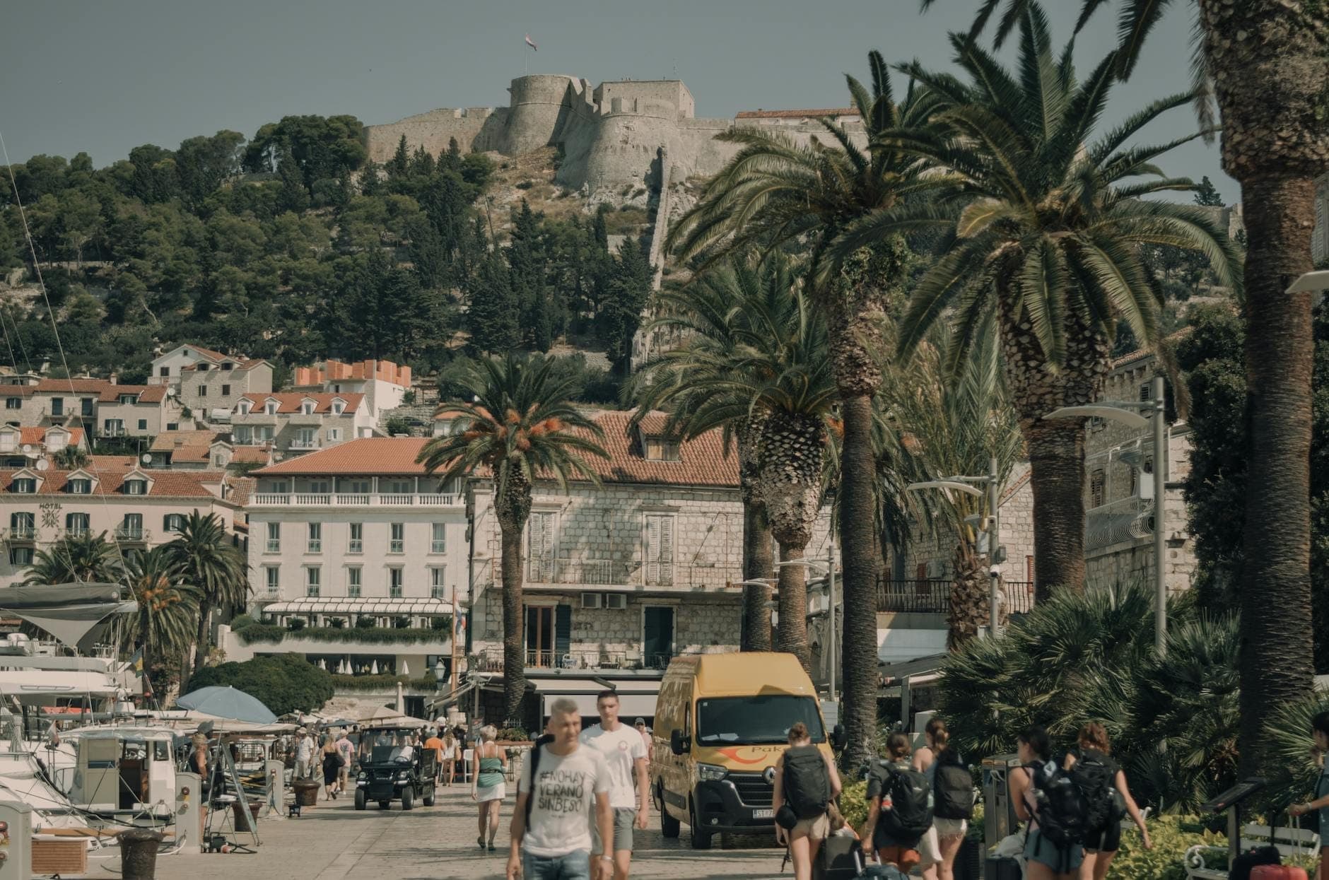 Pedoni che camminano lungo il lungomare del porto di Hvar con palme, edifici storici e la fortezza di Hvar sulla collina