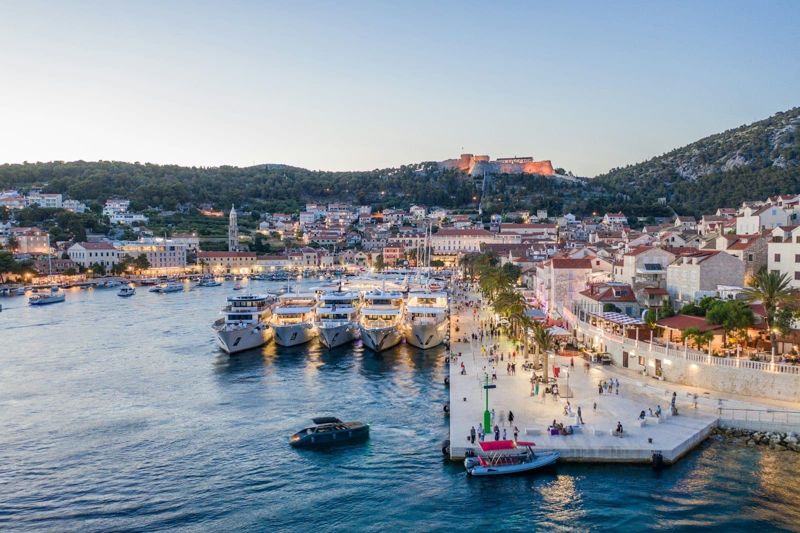 Il porto di Hvar al tramonto, con i traghetti attraccati sul lungomare, il centro storico sullo sfondo e le colline verdi che si alzano dietro la città.