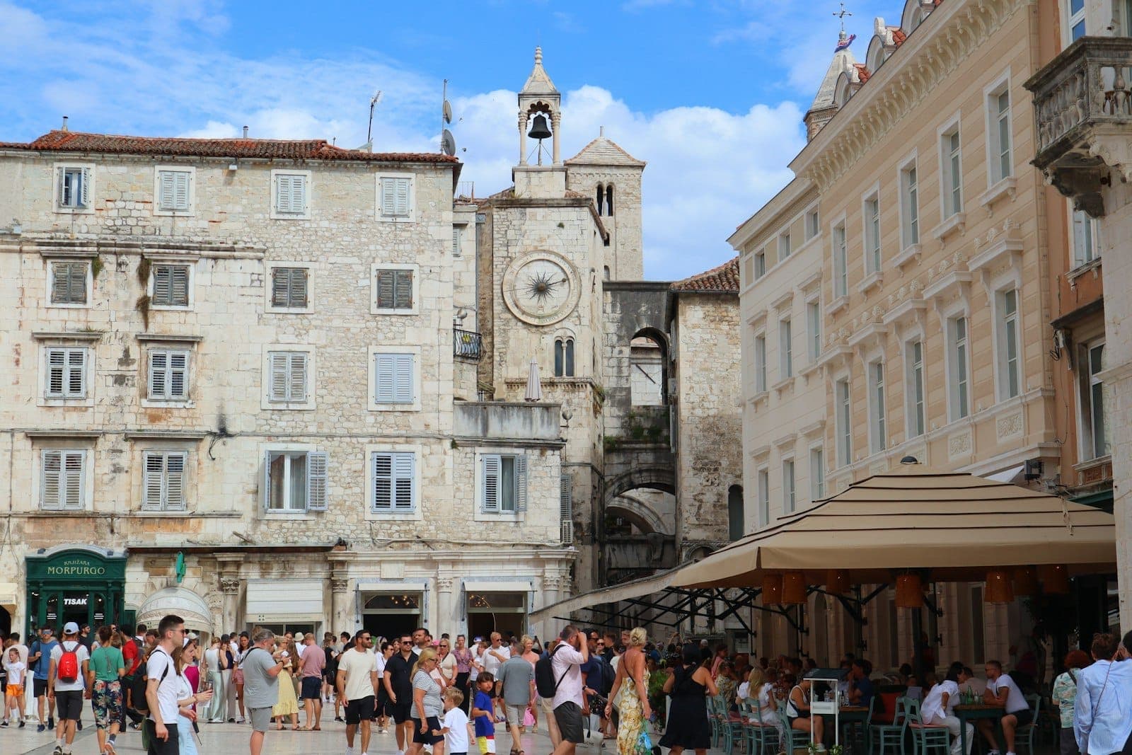 La piazza del centro storico di Spalato animata di turisti, con edifici in pietra, una torre dell'orologio e bar all'aperto sotto gli ombrelloni in una giornata di sole.