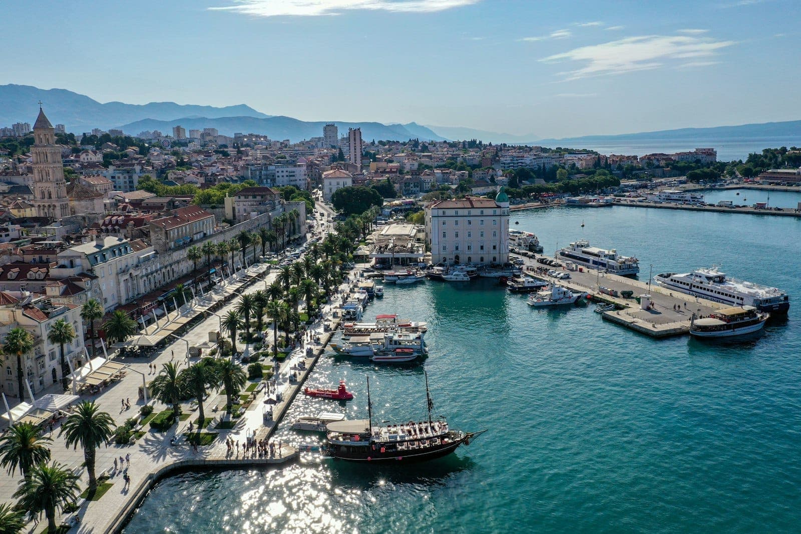 Vista aerea panoramica del porto di Spalato, con il lungomare alberato di palme, il centro storico e il limpido mare Adriatico color turchese sotto un cielo azzurro, con le montagne sullo sfondo.
