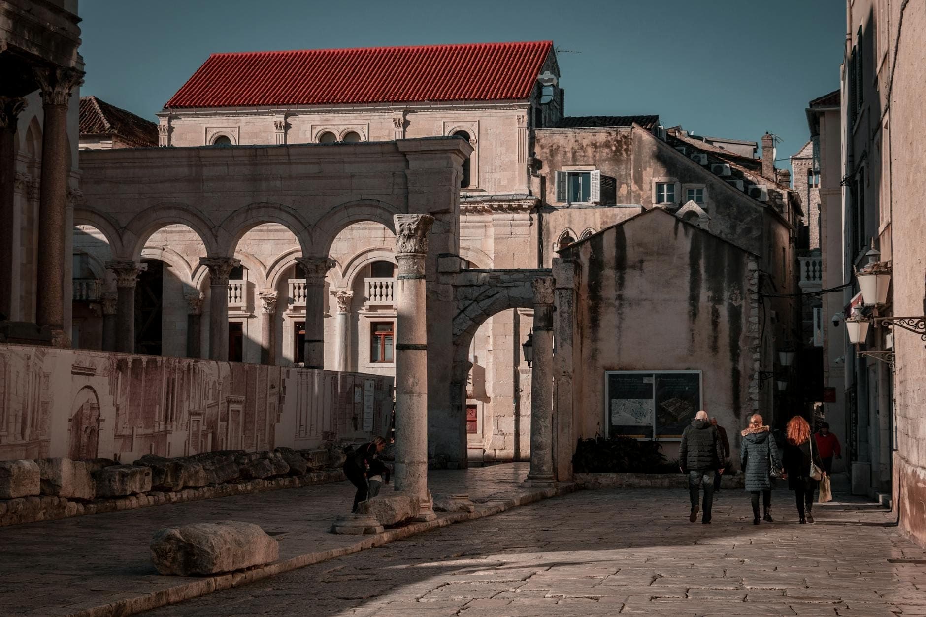 Persone che camminano vicino ad antichi archi romani e edifici in pietra nel centro storico di Spalato in una giornata di sole.
