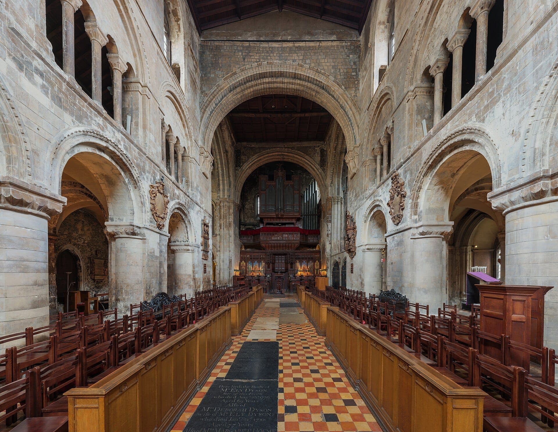 Wnętrze kościoła St Bartholomew the Great w Londynie z normańskimi łukami, kamiennymi kolumnami, drewnianymi ławkami i wzorzystą posadzką z płytek.