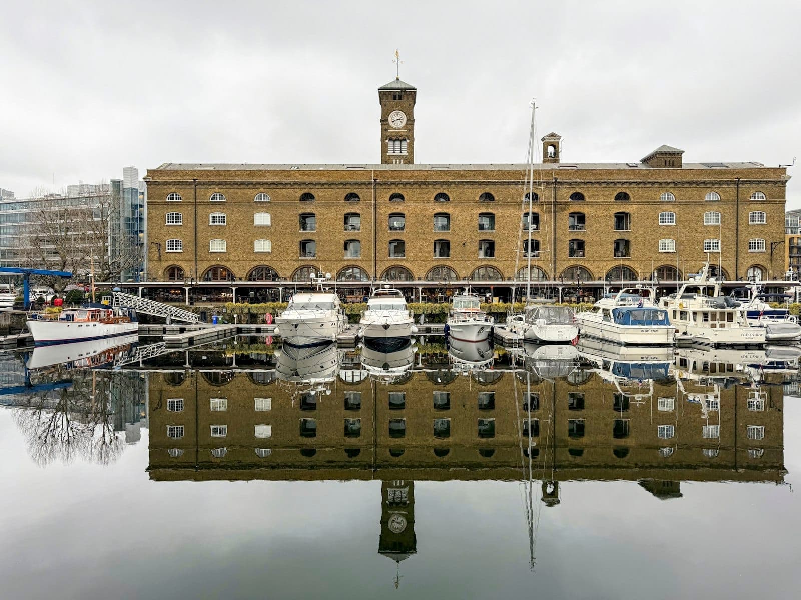 Marina de St Katharine Docks con histórico almacén de ladrillo, torre del reloj y barcos amarrados reflejados en aguas tranquilas en un día nublado.