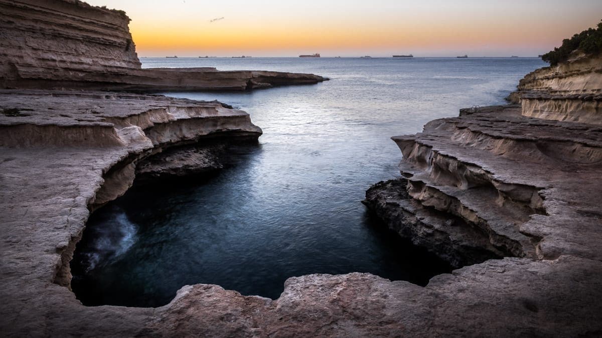 St. Peter's Pool, eine natürliche hufeisenförmige Kalksteinbucht an der Küste Maltas, mit geschichteten Felsvorsprüngen und ruhigem Meer bei Sonnenuntergang.