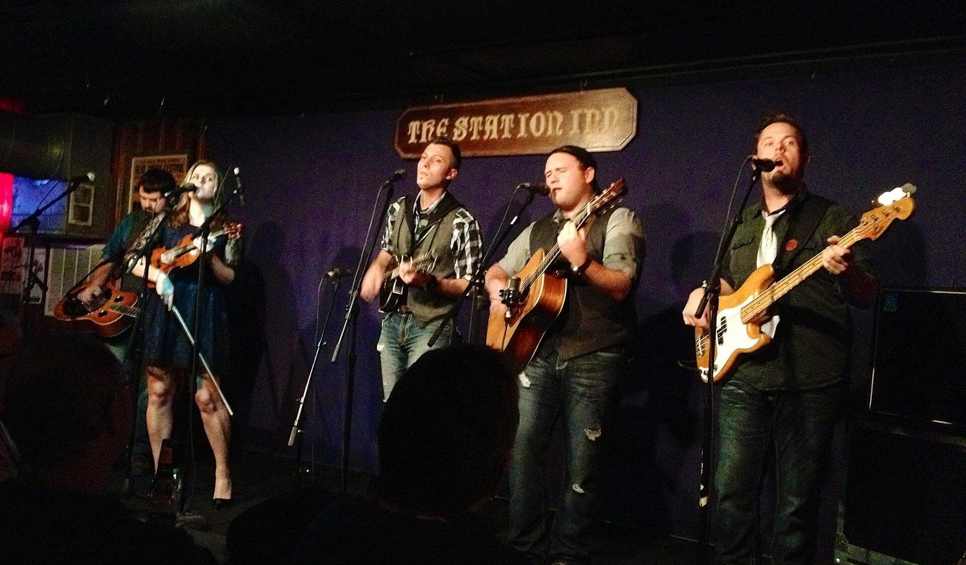 Five musicians perform with guitars, fiddle, and bass under The Station Inn sign on a dimly lit stage in Nashville.