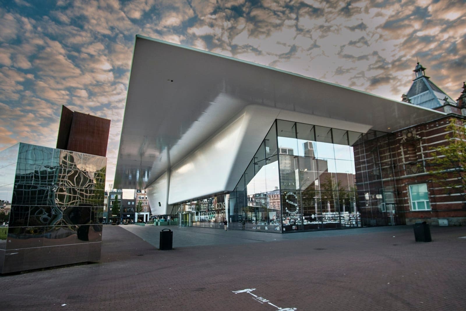 Moderne Glas- und Weißfassade des Stedelijk Museum Amsterdam unter einem dramatischen bewölkten Himmel, mit Spiegelungen in nahegelegenen Spiegelstrukturen.