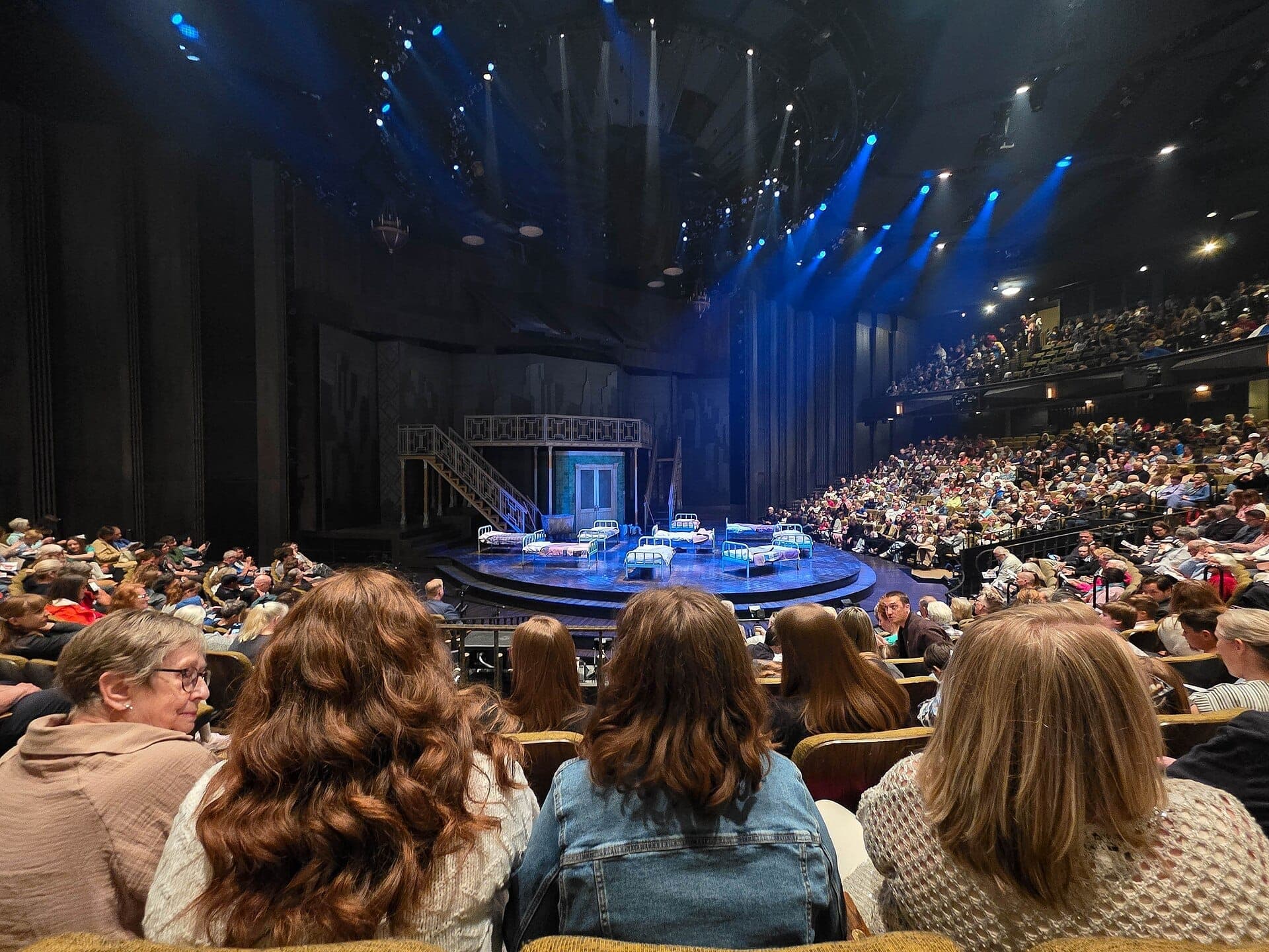 El público llena el teatro del Stratford Festival, frente a un escenario circular ambientado con camas e iluminación azul antes del inicio de una actuación.