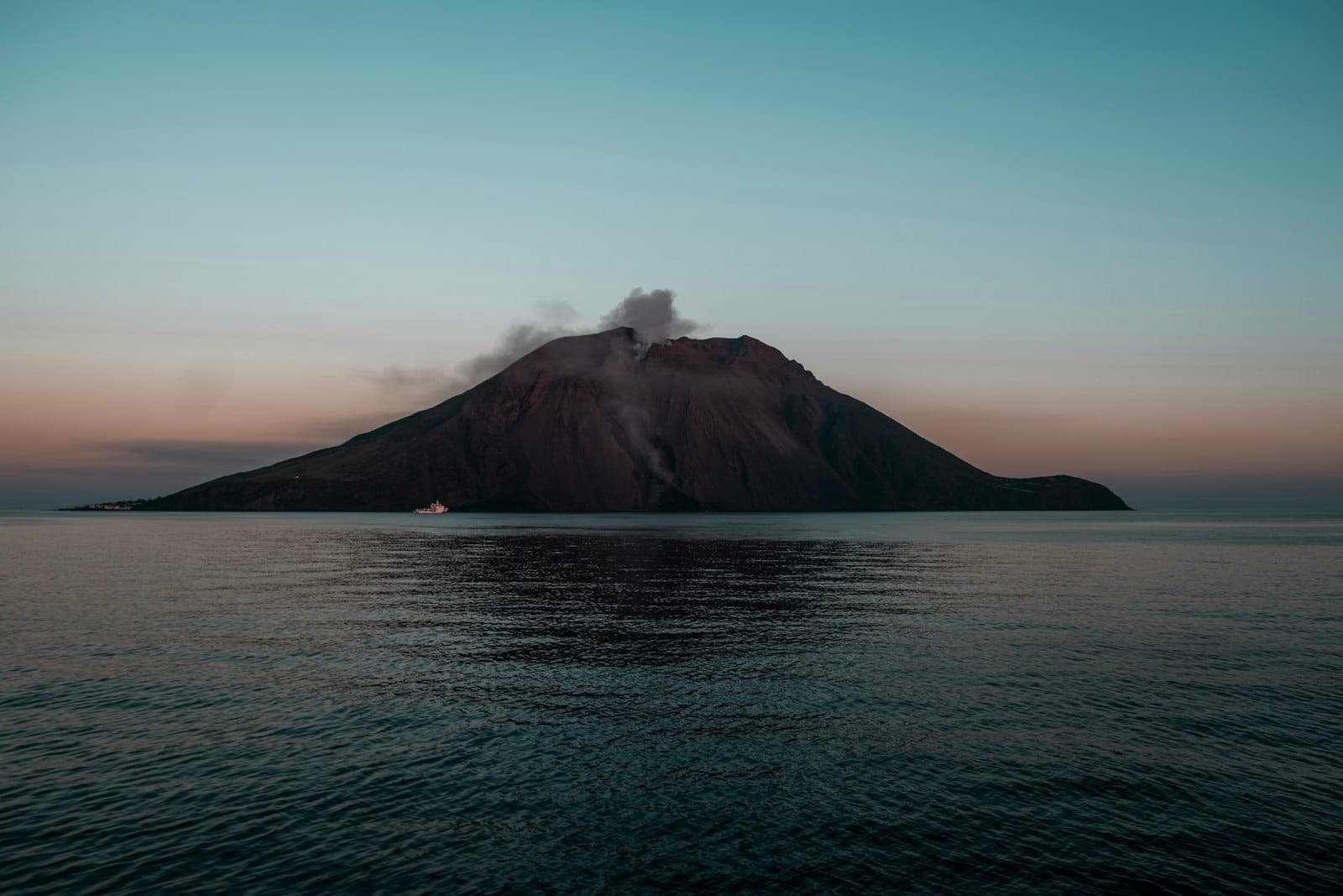 Gunung berapi Stromboli dengan asap mengepul dari kawahnya, terlihat melintasi perairan tenang saat senja dengan sebuah perahu di dekat tepian.