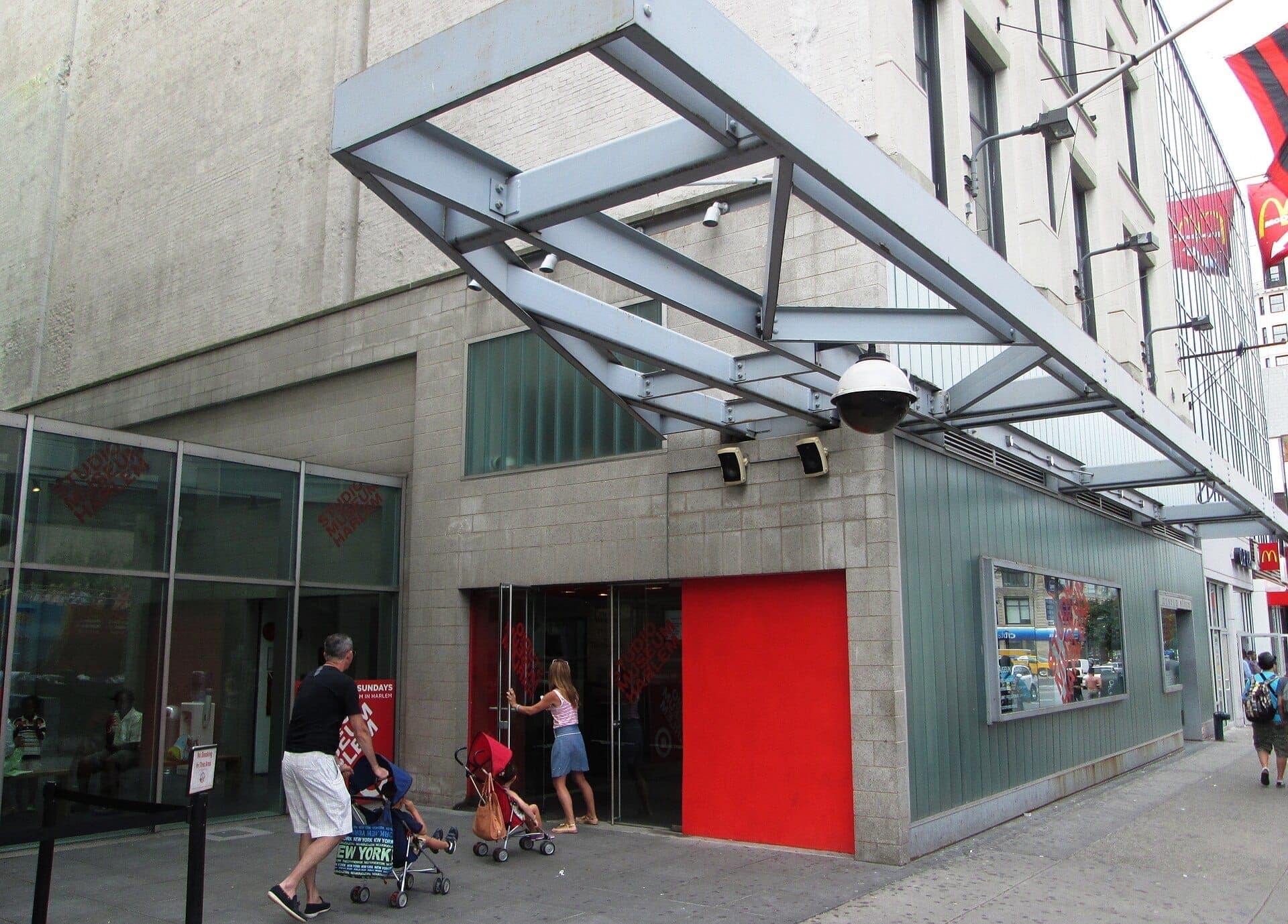 มุมมองภายนอกของ Studio Museum in Harlem โดดเด่นด้วยสถาปัตยกรรมสมัยใหม่ ผนังเน้นสีแดงสด และผู้คนที่กำลังเดินเข้าสู่อาคารบนทางเท้าที่คึกคัก