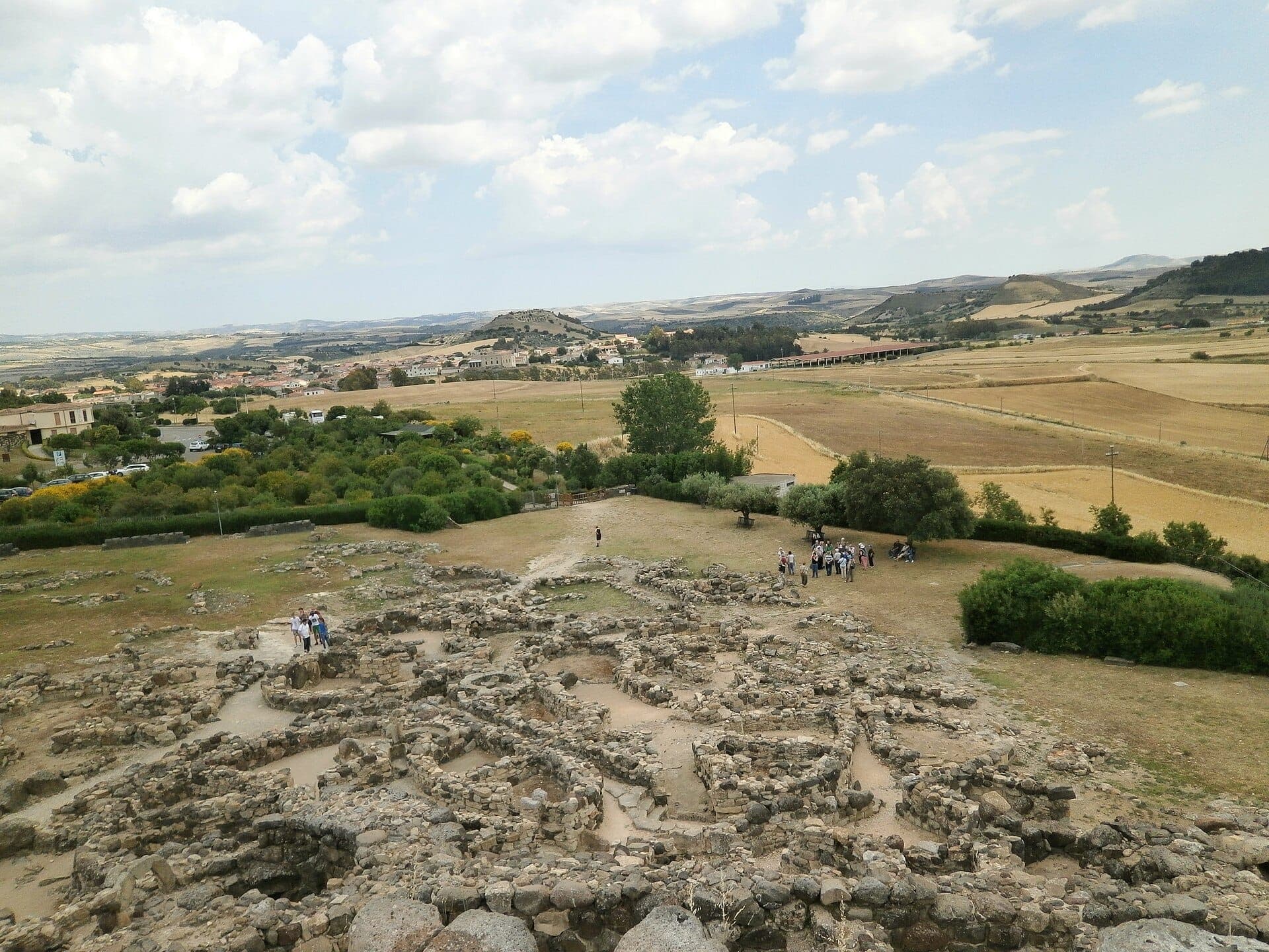 Ruinas de piedra de Su Nuraxi di Barumini extendidas por llanuras abiertas bajo un cielo nublado, con pequeños grupos de visitantes explorando el lugar.