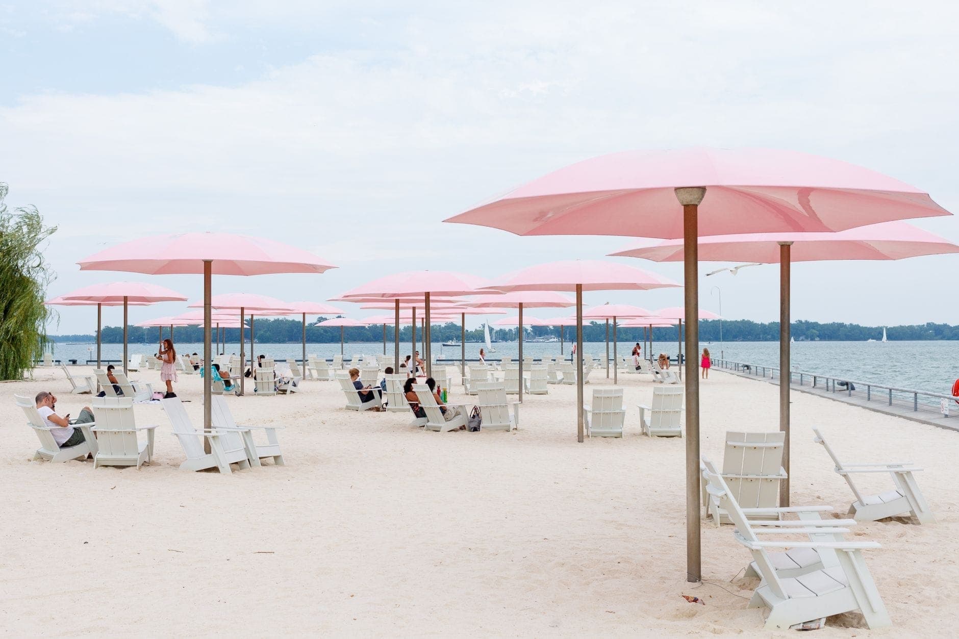 Una spiaggia urbana di Toronto con file di iconici ombrelloni rosa, sedie a sdraio bianche e persone che si rilassano sul lungofiume, con il lago Ontario sullo sfondo.