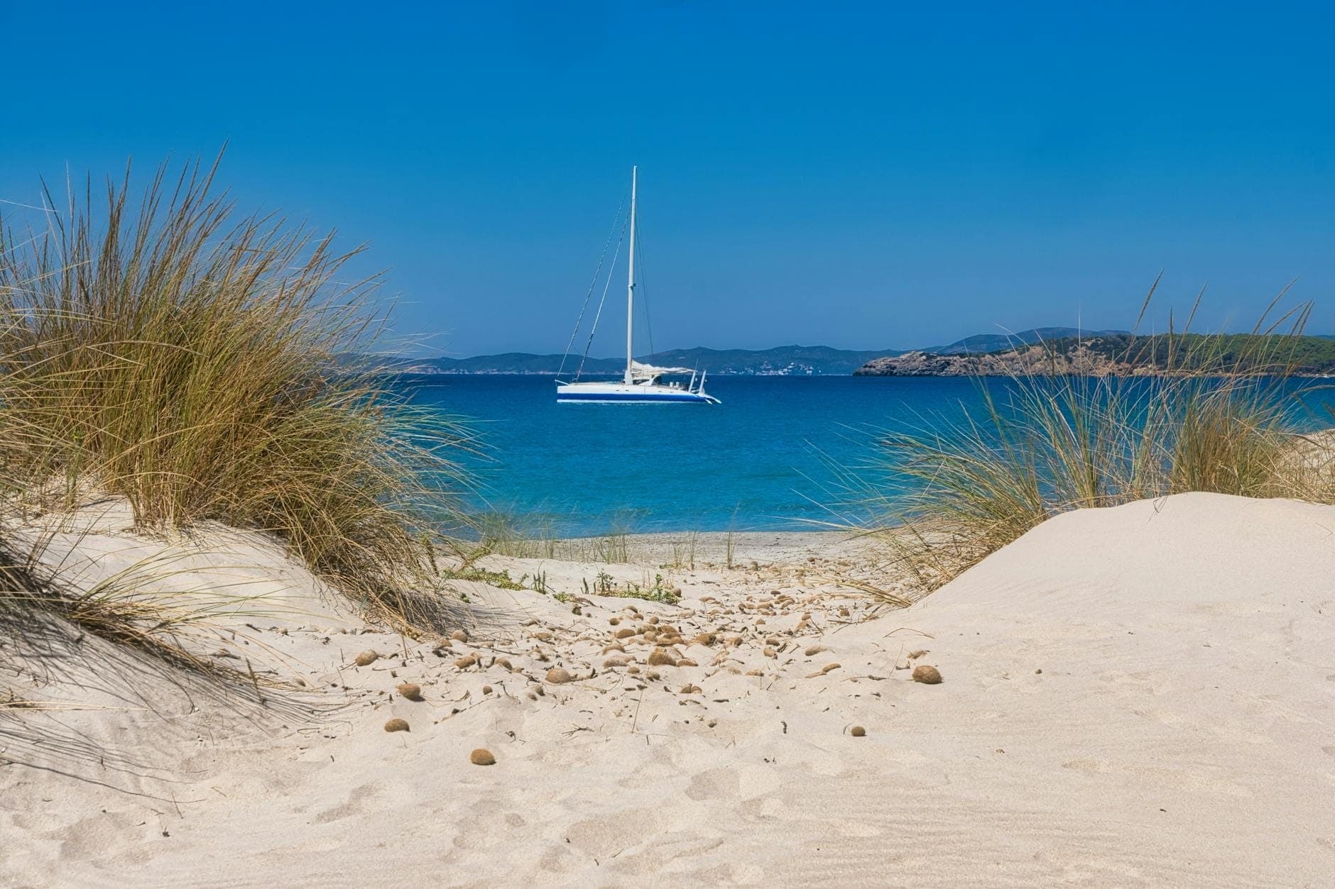 Dunas de arena blanca con hierbas secas en Porto Pino, Sulcis, con vistas a un catamarán en el claro mar Mediterráneo azul.