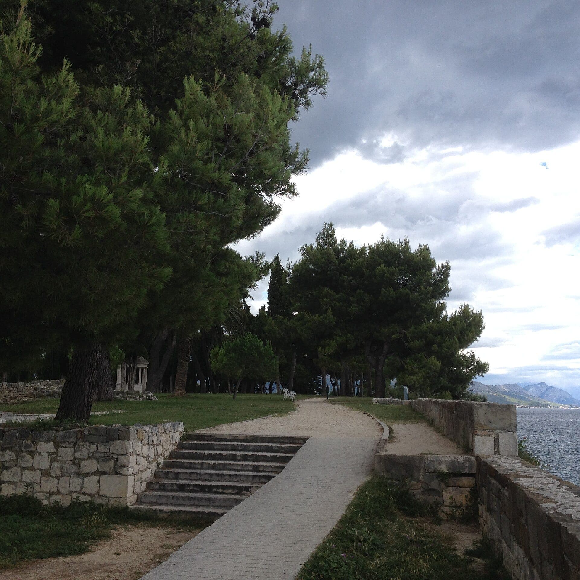 Un sentier en pierre et des marches traversent de luxuriants pins sur Sustipan, surplombant la mer Adriatique et des montagnes lointaines sous un ciel nuageux.