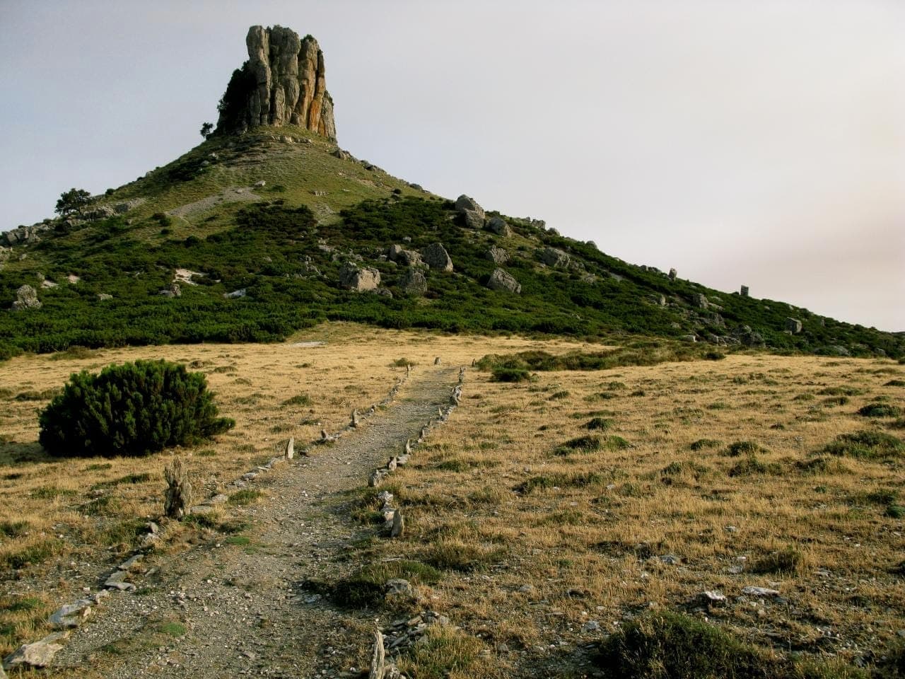 Un escarpado afloramiento de piedra caliza de Tacchi d'Ogliastra se eleva sobre llanuras herbosas y arbustos bajos bajo un cielo pálido y amplio.