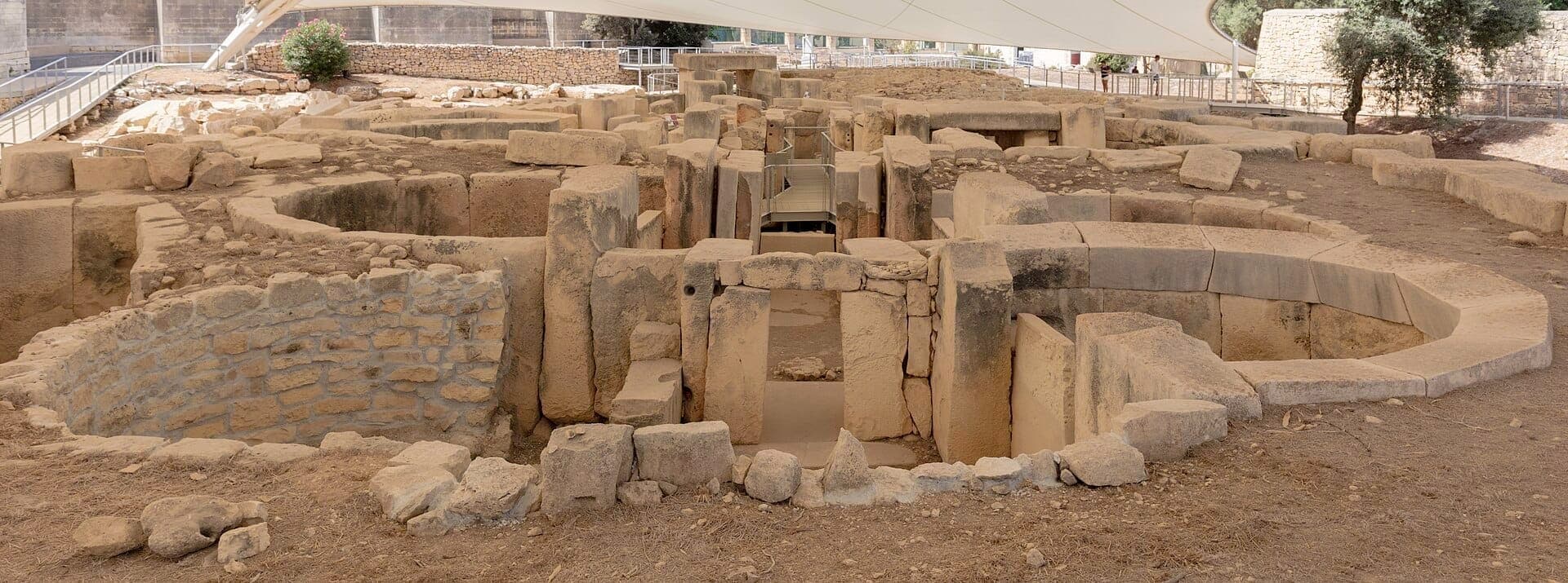 Die prähistorischen Ruinen der Tarxien Temples mit großen verwitterten Steinblöcken und kreisförmigen Gehegen unter einem Schutzdach mit Wegen im Hintergrund.