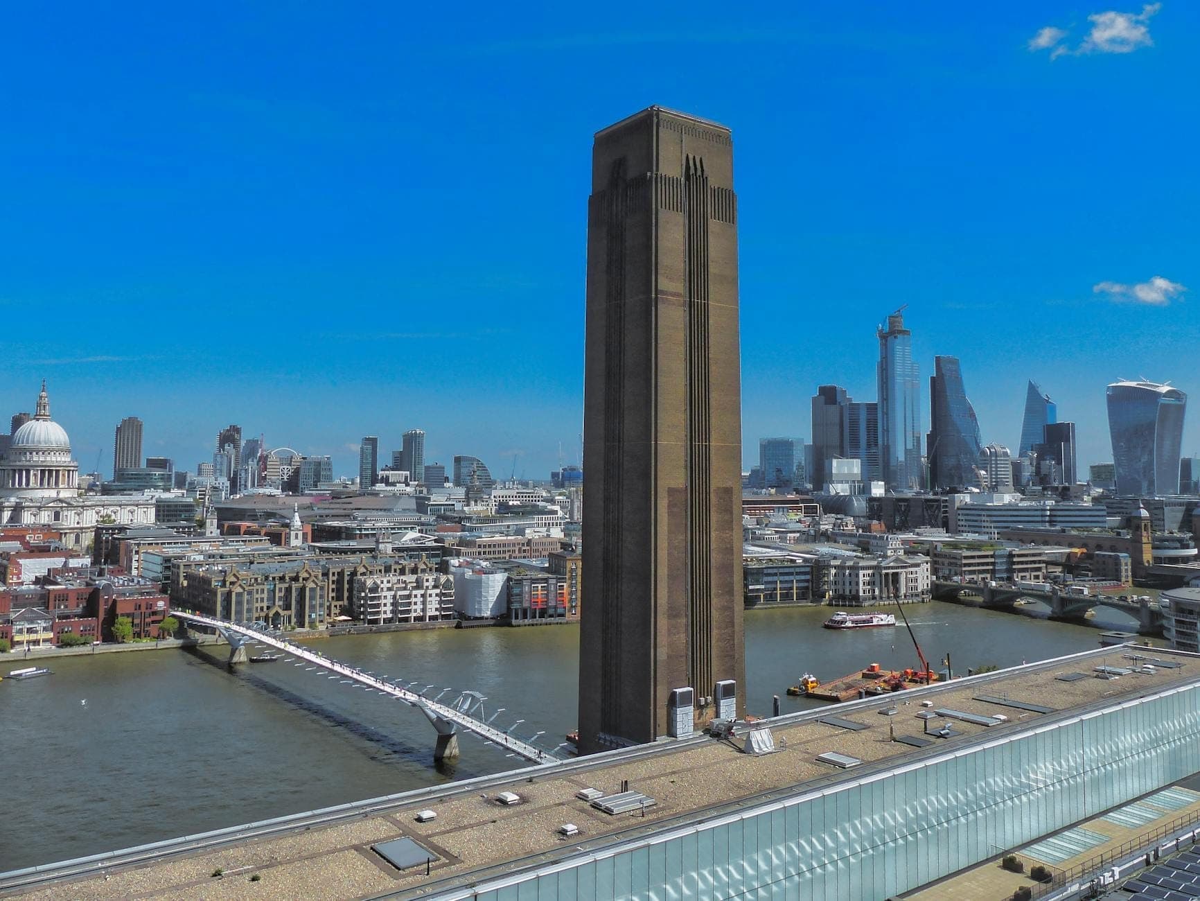 Vue de la cheminée emblématique de la Tate Modern avec la Tamise, le Millennium Bridge et le skyline de Londres sous un ciel bleu éclatant.