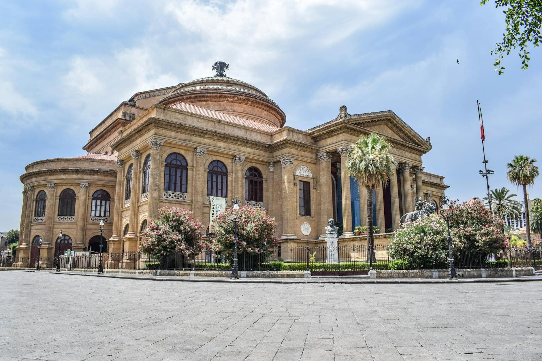 Tampak lebar gedung opera Teatro Massimo bergaya neoklasik di Palermo, diapit pohon palem dan dedaunan warna-warni di bawah langit biru cerah.