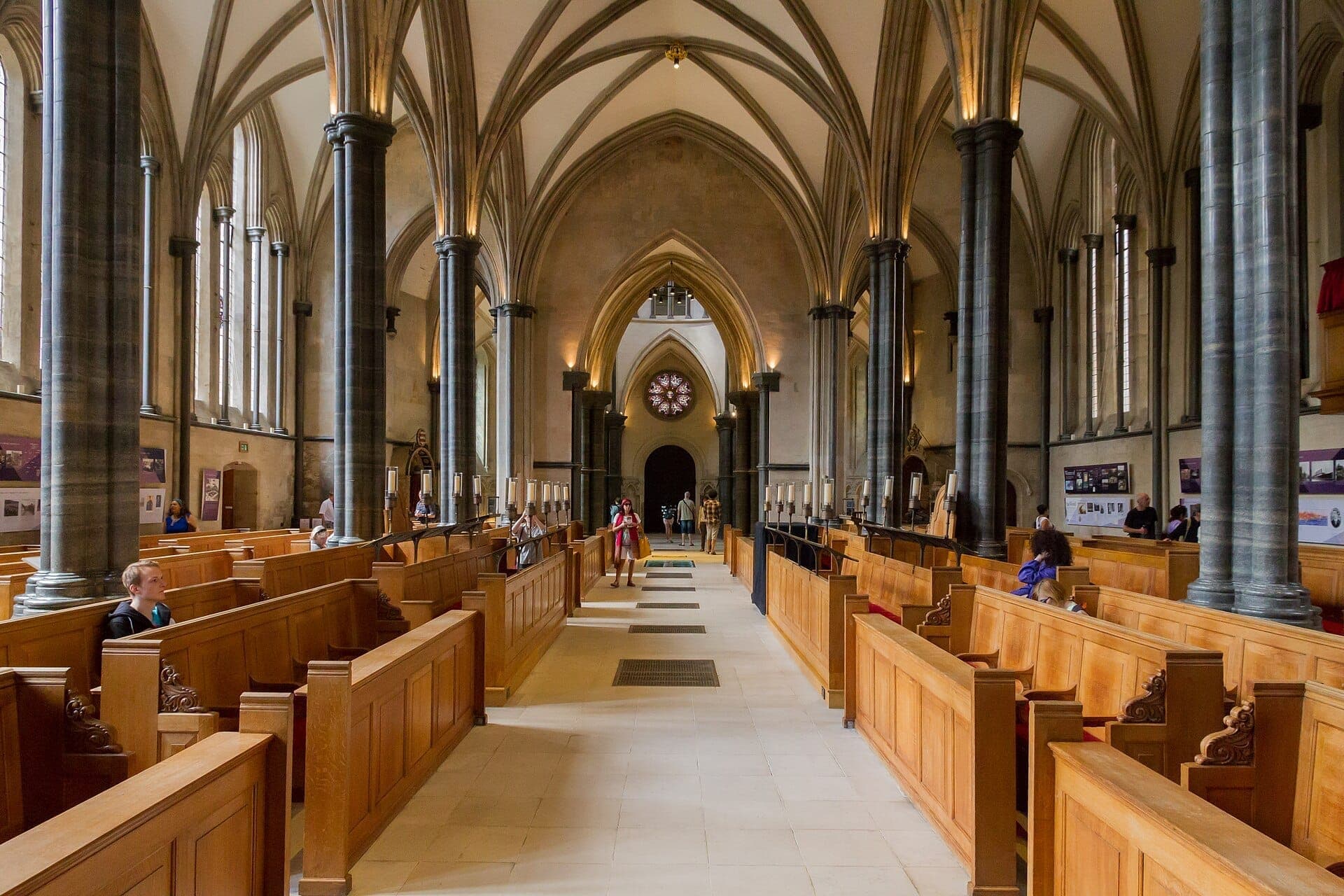 Widok wnętrza Temple Church w Londynie z wysokimi gotyckimi łukami, kamiennymi kolumnami, drewnianymi ławkami, witrażami i kilkoma zwiedzającymi.