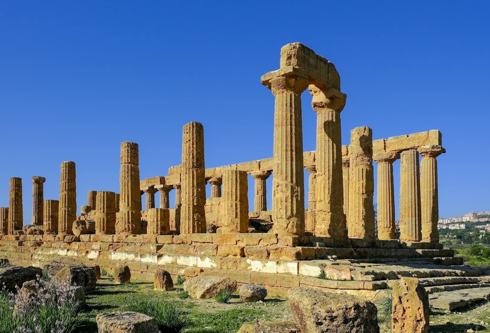 Kuil Juno di Agrigento berdiri megah di atas perbukitan berbatu, dengan tiang-tiang berwarna madu berlatar langit biru Mediterania yang cerah.