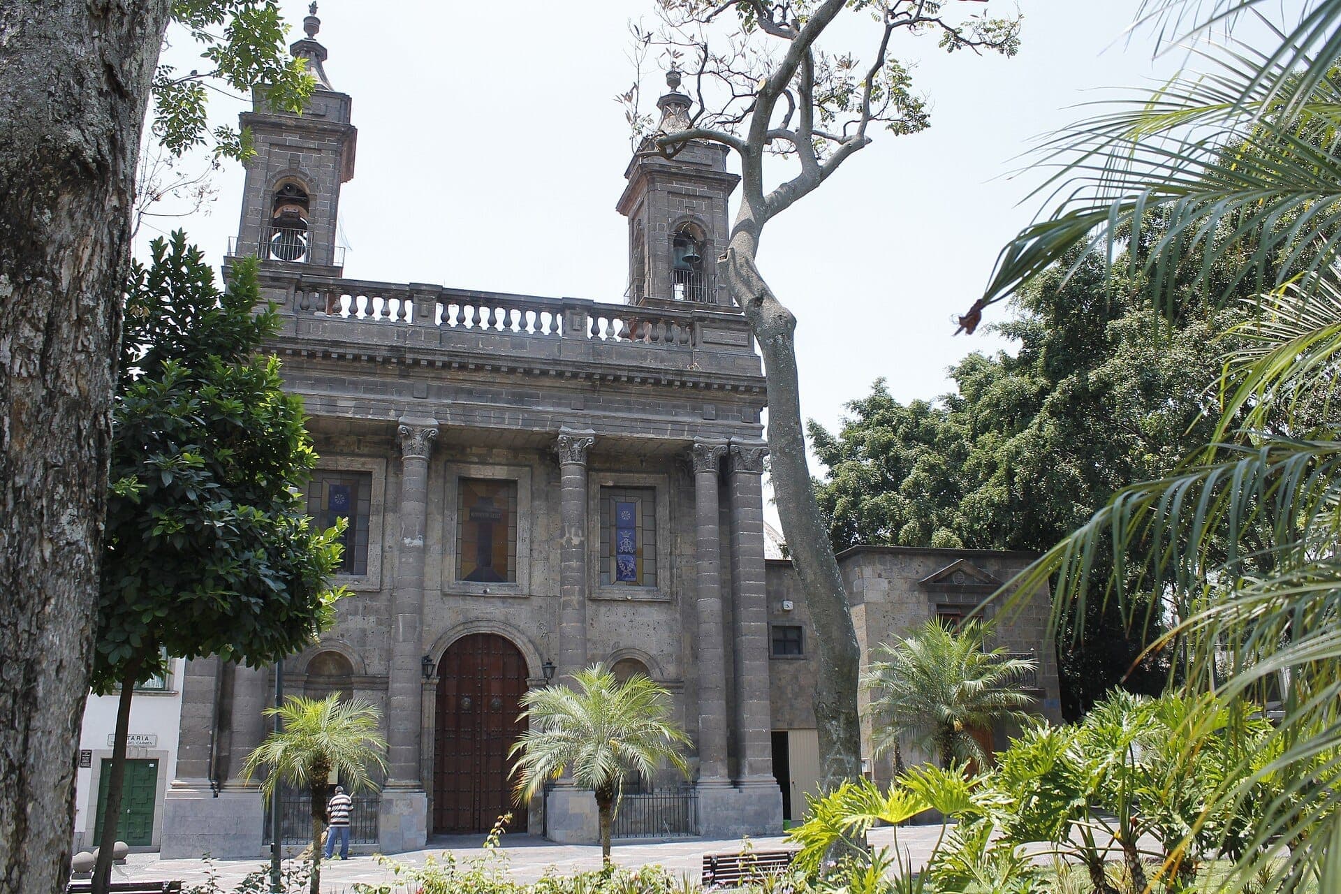 มุมมองด้านหน้าของ Templo del Carmen โบสถ์หินสไตล์นีโอคลาสสิกที่ล้อมรอบด้วยต้นปาล์มและต้นไม้สีเขียวในใจกลางเมืองเก่าของ Guadalajara