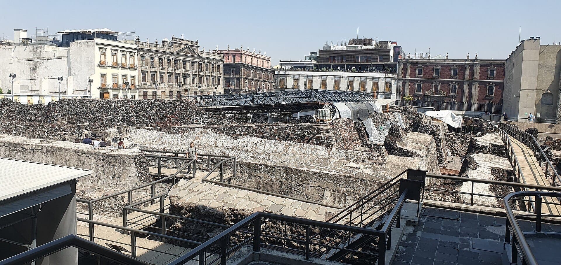 ซากปรักหักพังหินของ Templo Mayor พร้อมทางเดินและนักท่องเที่ยว ล้อมรอบด้วยอาคารประวัติศาสตร์ใต้ท้องฟ้าสีฟ้าใสใน Mexico City