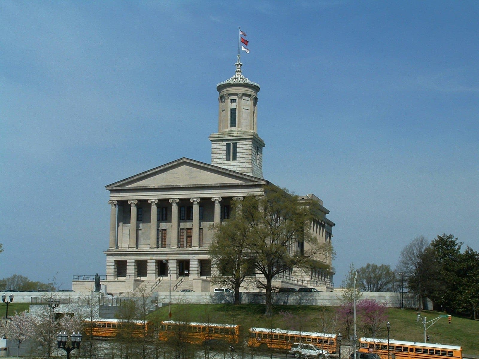Landscape view of the Tennessee State Capitol building under a blue sky with green trees and yellow school buses in the foreground.