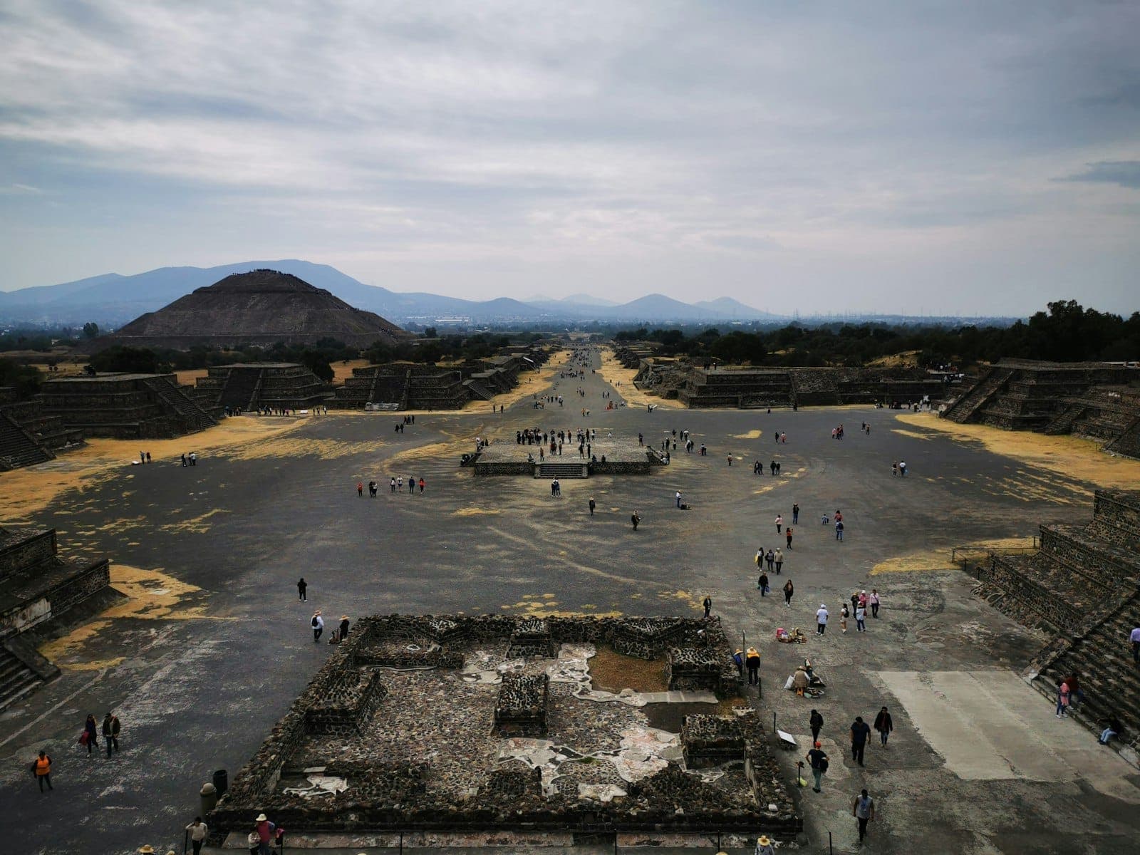 Vue le long de l'Avenue des Morts à Teotihuacán, avec des visiteurs se promenant parmi les ruines anciennes et la Pyramide de la Lune au loin.