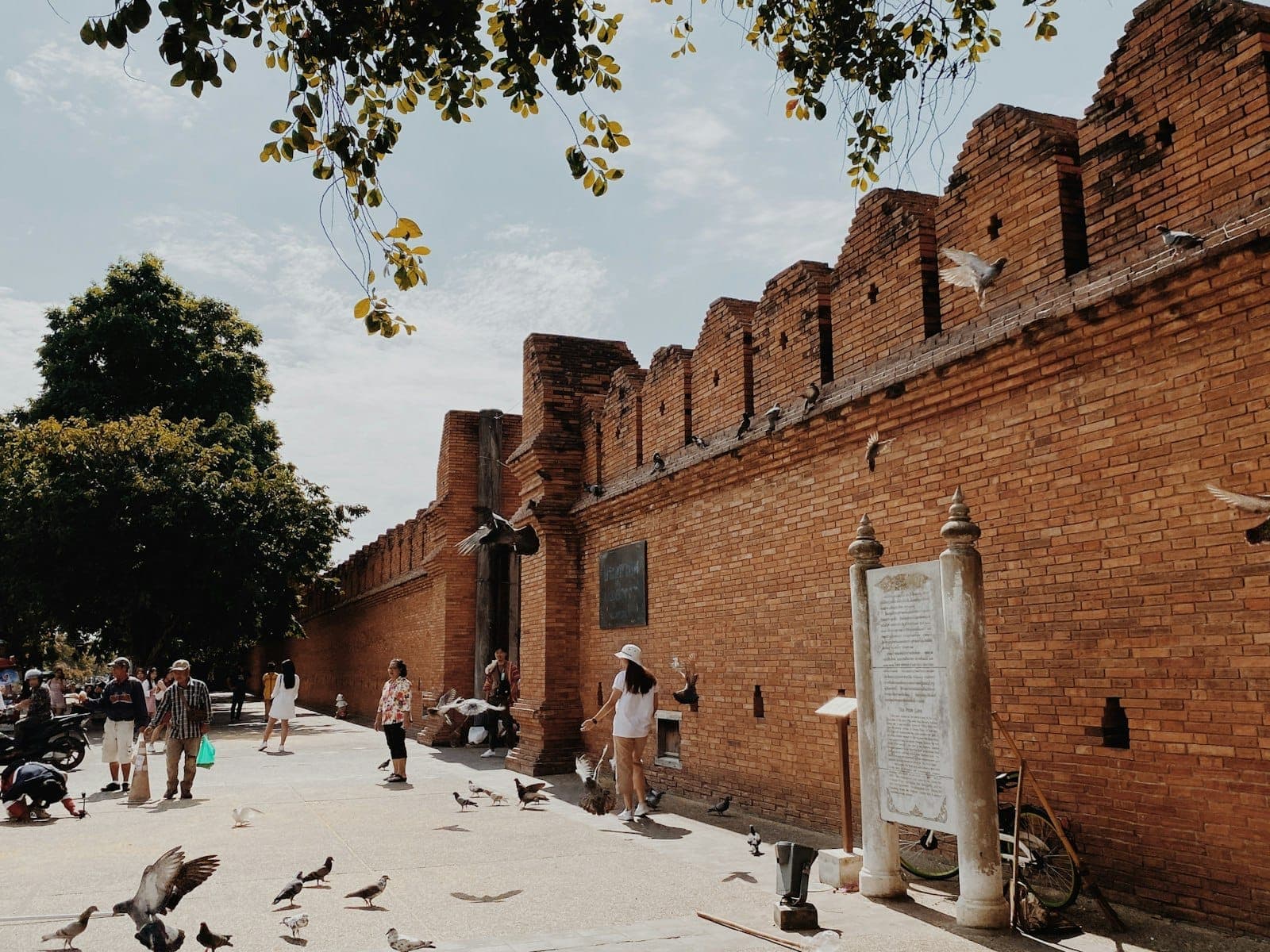 Visitors walk and feed pigeons near the iconic brick walls of Tha Phae Gate in Chiang Mai under a partly cloudy sky.