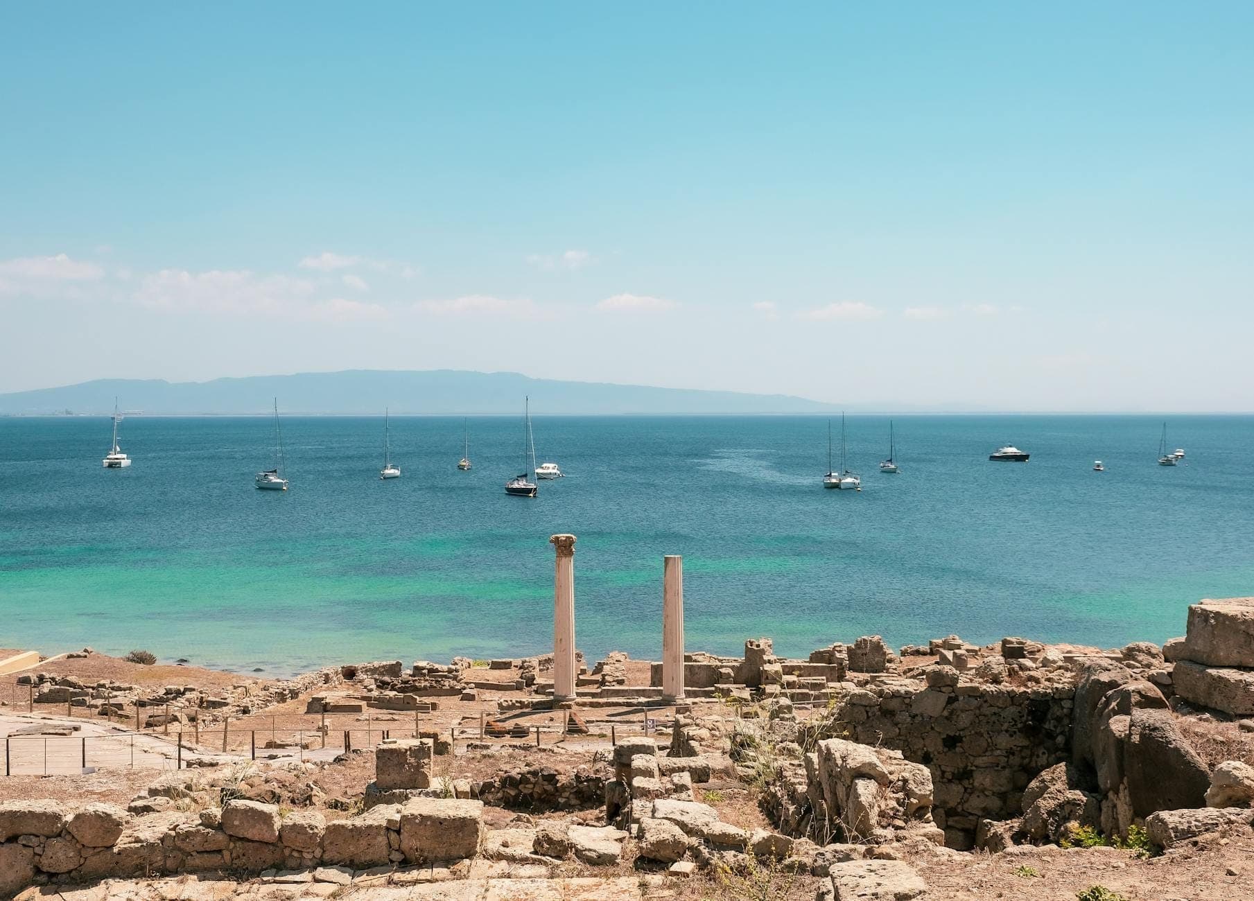 Ruinas de piedra antigua con dos columnas romanas en pie que dan al mar turquesa, con veleros a lo lejos y un cielo azul intenso.