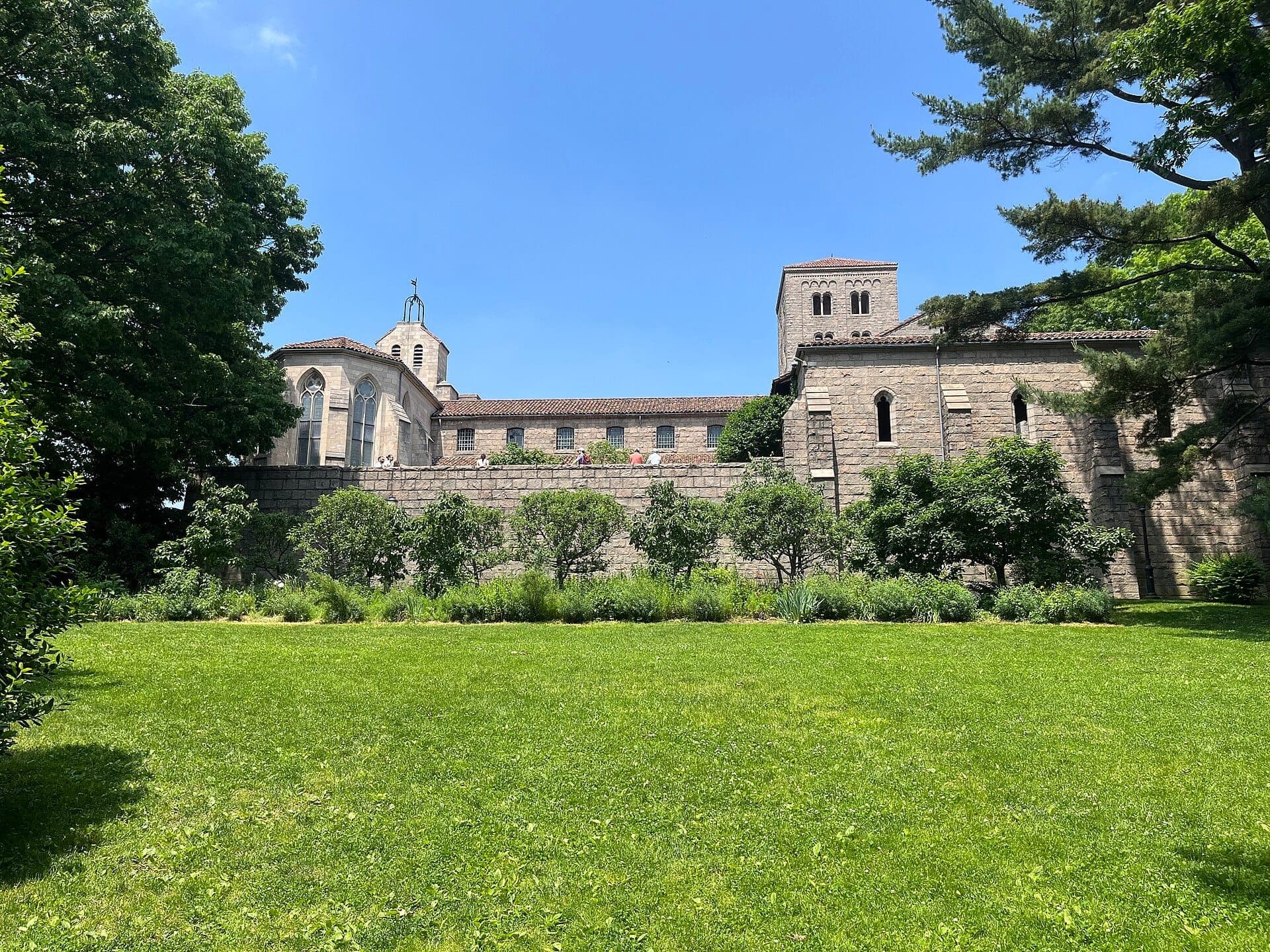 วิวภายนอกของ The Met Cloisters พร้อมกำแพงหินสมัยกลาง หอคอย สวนภูมิทัศน์ และต้นไม้เขียวขจีใต้ท้องฟ้าสีฟ้าสดใส